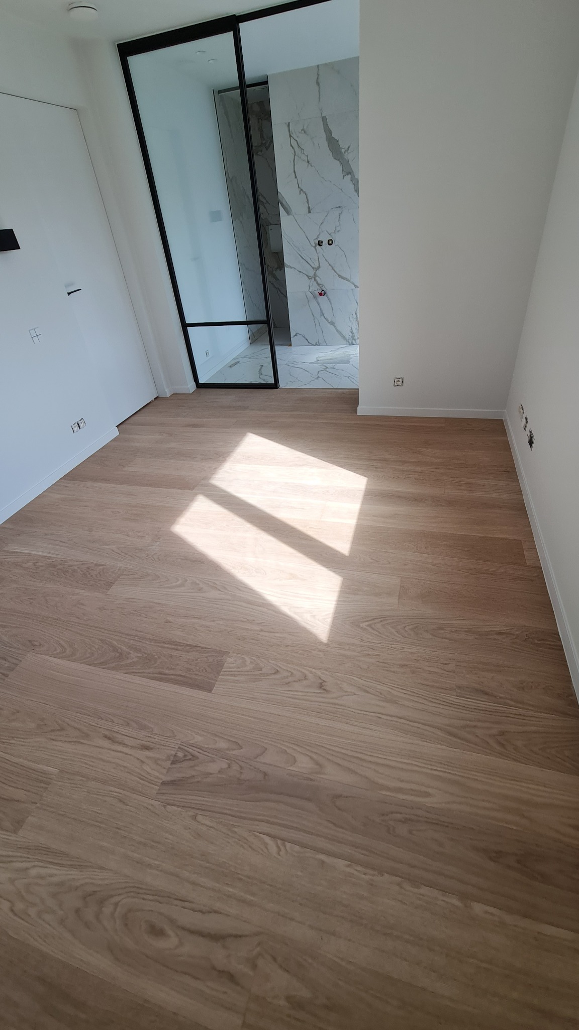 Jasne, drewnopodobne panele podłogowe w nowym pomieszczeniu, widoczny fragment łazienki z marmurowymi płytkami, wpadające promienie słoneczne.