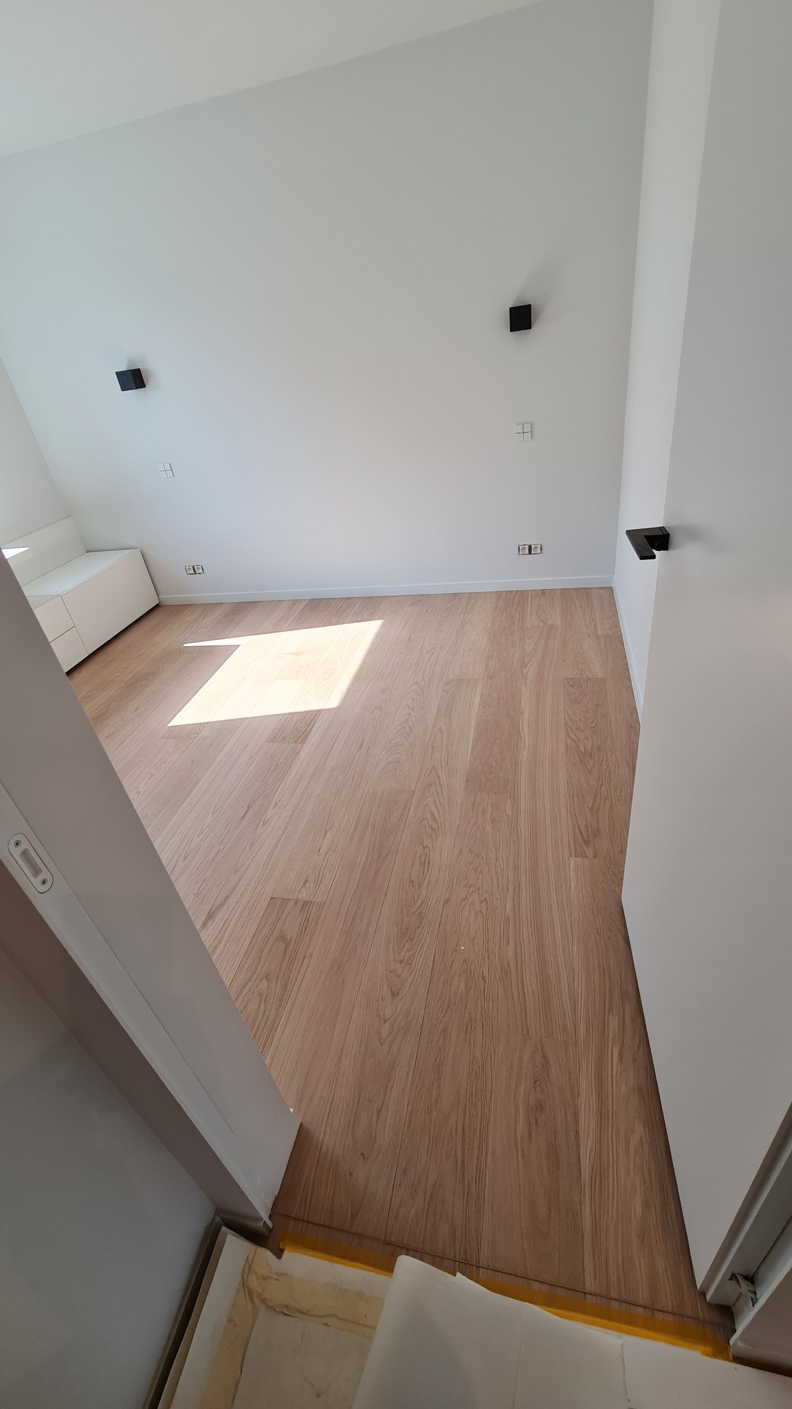 Jasne panele podłogowe w minimalistycznym wnętrzu, widoczne światło słoneczne, czarne punkty oświetleniowe na białej ścianie, białe meble w tle, kadr z perspektywy wejścia.