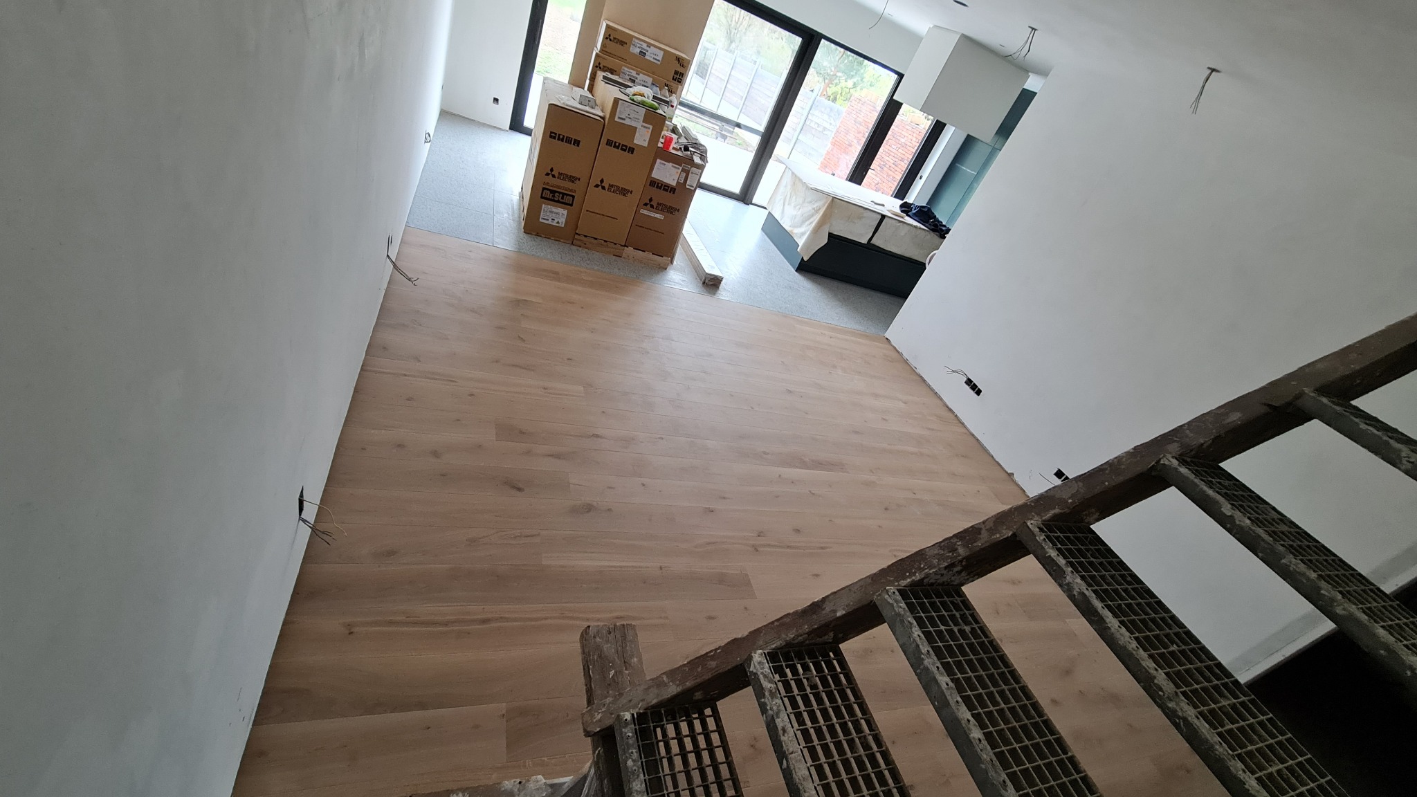 Jasne, drewniane deski podłogowe ułożone w przestronnym, surowym wnętrzu. Widoczne kartony z urządzeniami i fragment metalowych schodów w kadrze.