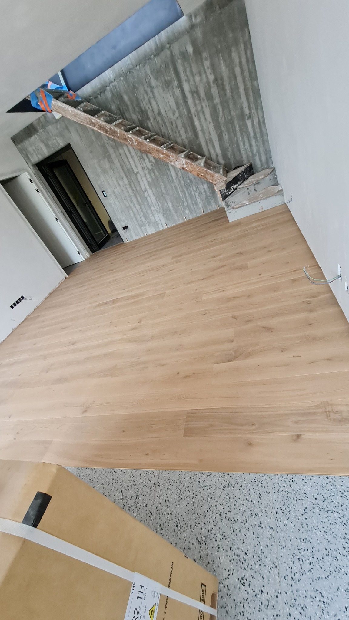 Jasne panele podłogowe ułożone w pokoju z surowymi betonowymi ścianami i schodami, widoczna kartonowa paczka paneli w rogu.