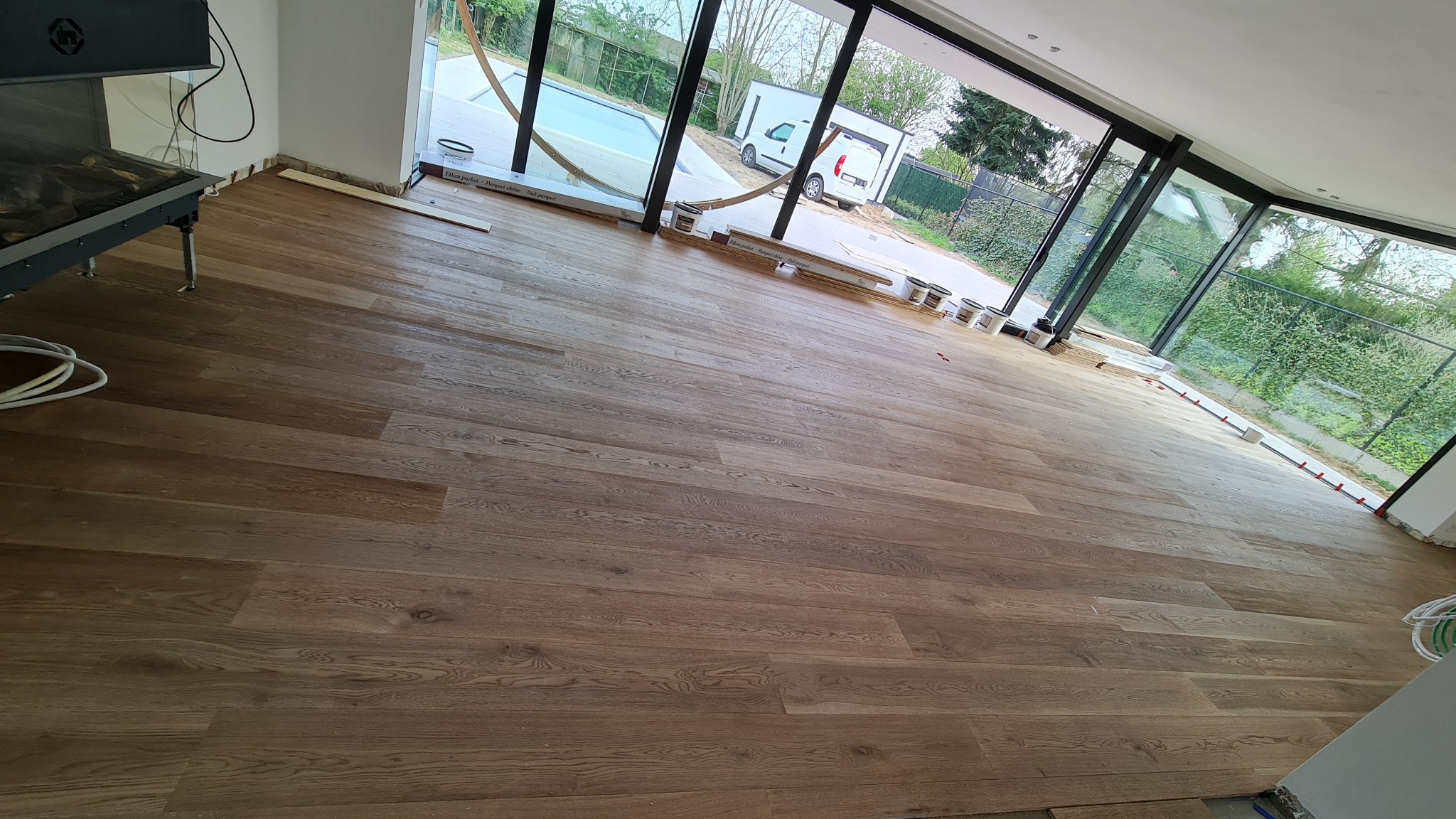 Nowo ułożona, lakierowana podłoga z desek w jasnym odcieniu brązu, widoczne paczki z panelami przy oknie i akcesoria montażowe, duże okna z widokiem na basen i zieleń.