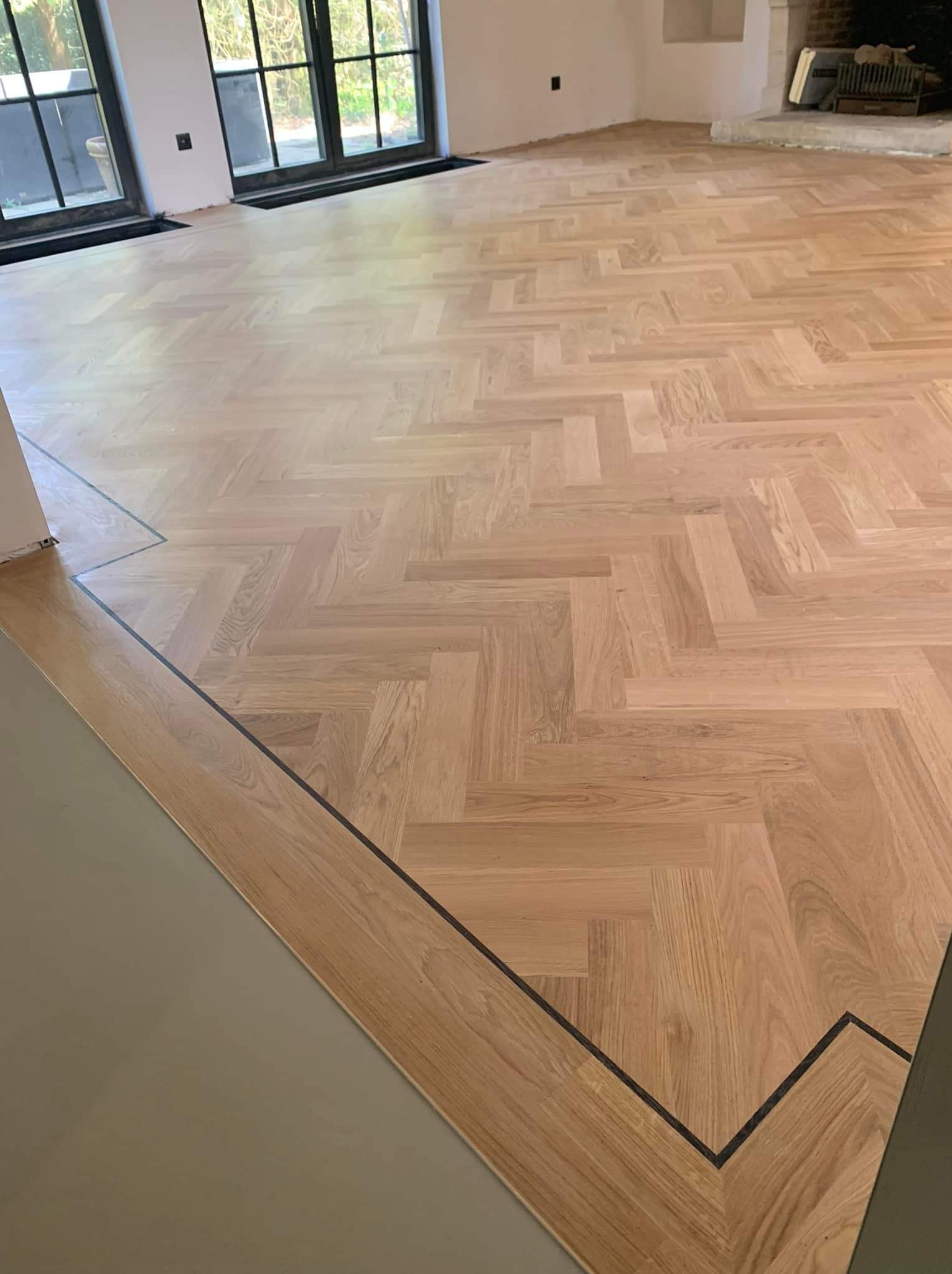 Nowo położony, jasny parkiet ułożony we wzór jodełki francuskiej z ciemną, cienką linią obramowania, widoczne okna i kominek w tle.