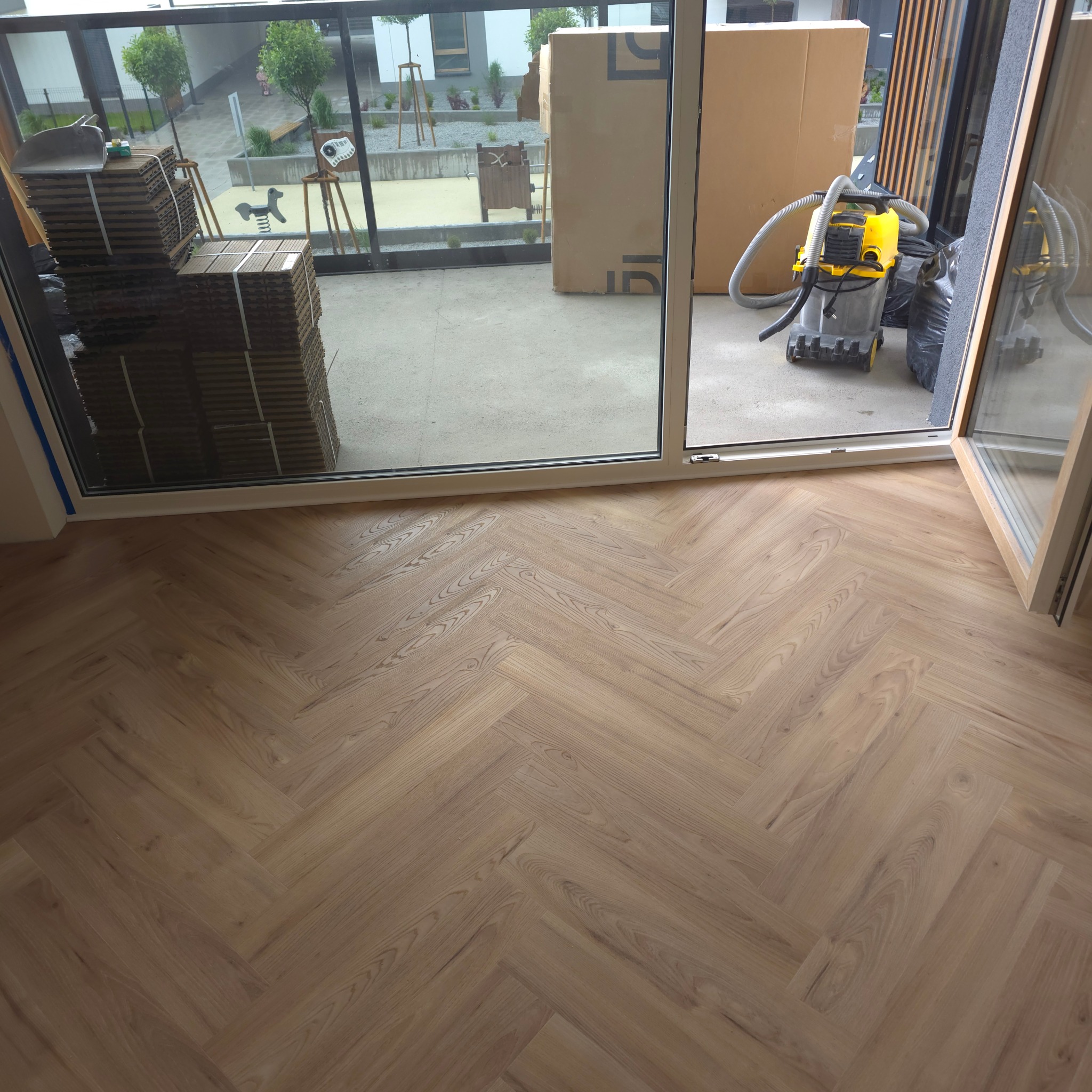 Ułożony parkiet w jodełkę na balkonie, z widocznymi paczkami paneli i odkurzaczem przemysłowym w tle. Jasne drewno, naturalne światło.