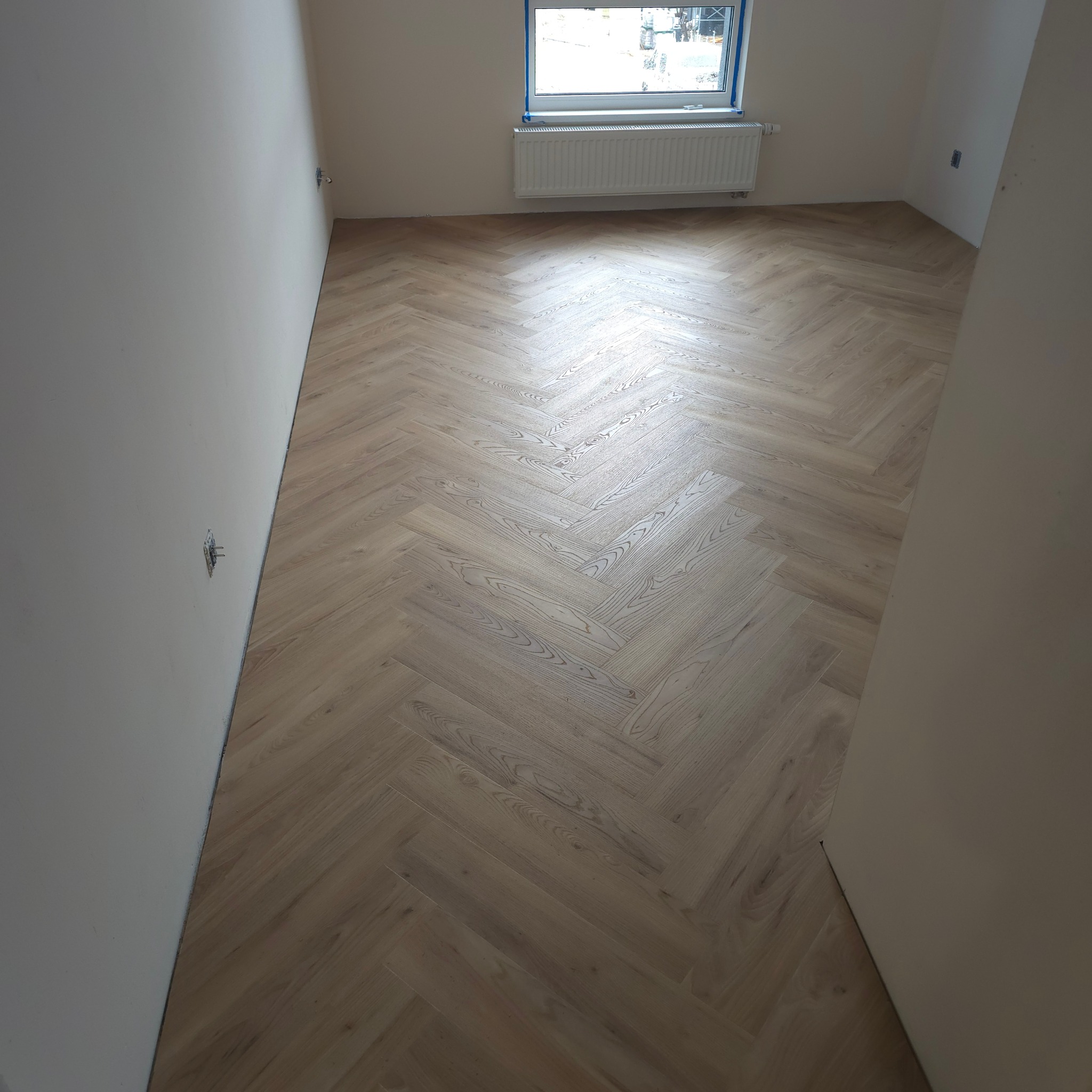 Jasne parkiet ułożony w jodełkę klasyczną w pustym pokoju z oknem i grzejnikiem. Widoczne gniazdka elektryczne na ścianie.