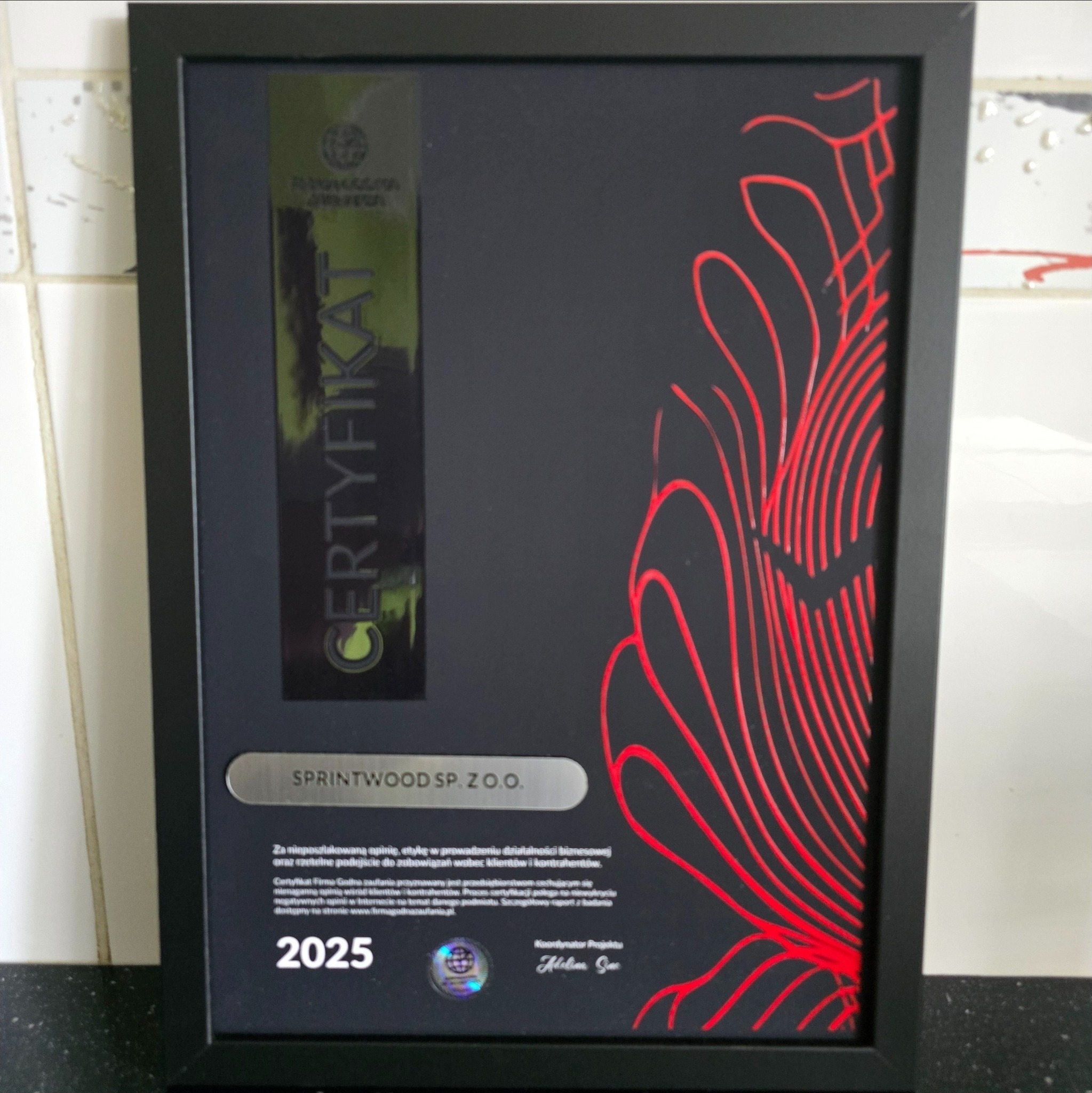 Oprawiony certyfikat Sprintwood Sp. z o.o. z czerwonymi zdobieniami, przyznany za etykę i rzetelność w biznesie, ważny do 2025 roku. Detaliczny widok, czarna ramka.