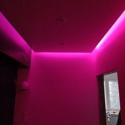 CORBUD - Wnętrze z podświetleniem LED w kolorze fuksji, widoczne fragmenty sufitu podwieszanego, ściany pomalowane na jasny kolor, gniazdko elektryczne oraz fragment futryny drzwiowej z widocznym oknem...