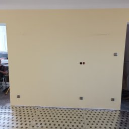 CORBUD - Ściana w trakcie przygotowywania do malowania, widoczne puszki elektryczne i zabezpieczona folią podłoga z nadrukiem, fragment sylwetki osoby w tle.