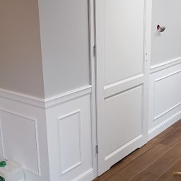 CORBUD - Białe, dwuskrzydłowe drzwi wewnętrzne zamontowane w ścianie z dekoracyjną sztukaterią, widoczne brązowe panele podłogowe i pojemnik z płynem do czyszczenia w rogu.