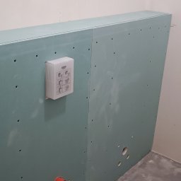 CORBUD - Stelaż z płyty karton-gips w łazience, widoczne otwory na instalacje, zamontowana puszka Grohe ze schematem montażu, surowy beton na podłodze.