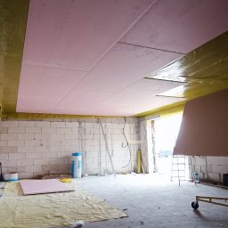 CORBUD - Montaż płyt kartonowo-gipsowych na suficie w surowym pomieszczeniu, widoczne profile, izolacja i przygotowane płyty na podłodze.