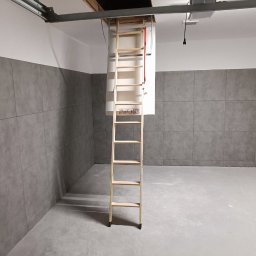 CORBUD - Zamontowane drewniane schody strychowe prowadzące do otworu w suficie w pomieszczeniu z szarymi płytkami na ścianie i betonową podłogą.