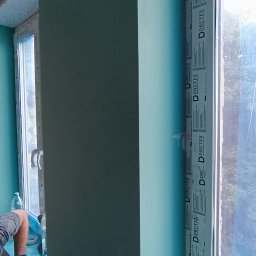 CORBUD - Montaż pionowej konstrukcji z płyty gipsowo-kartonowej przy oknie z widocznym logo producenta, fragment pracownika w tle, zabezpieczona folią podłoga.