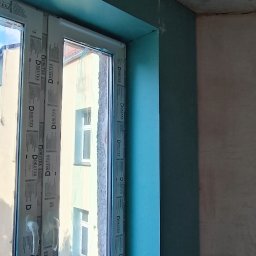 CORBUD - Montaż zielonej płyty kartonowo-gipsowej wokół okna, widoczna wiertarka akumulatorowa na parapecie, surowy sufit w tle.