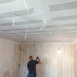 CORBUD - Mężczyzna w krótkich spodenkach i czarnej koszulce maluje ścianę wałkiem w pomieszczeniu z płytami kartonowo-gipsowymi na suficie, widoczne przewody elektryczne, wiaderka z farbą i odkurzacz...