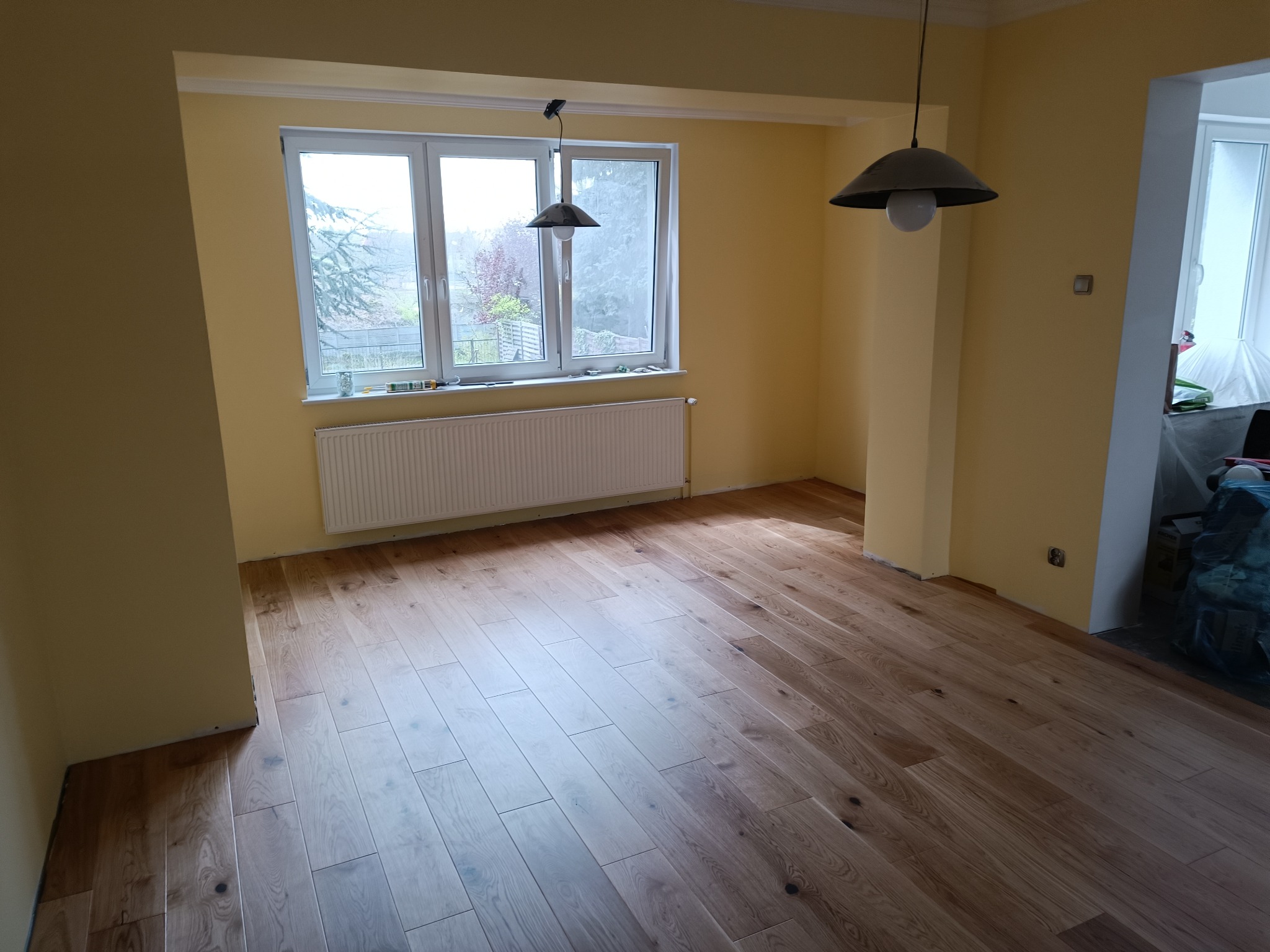 Jasne, drewniane panele podłogowe ułożone w pustym pokoju z żółtymi ścianami, białym grzejnikiem pod oknem i dwoma lampami wiszącymi w stylu retro.