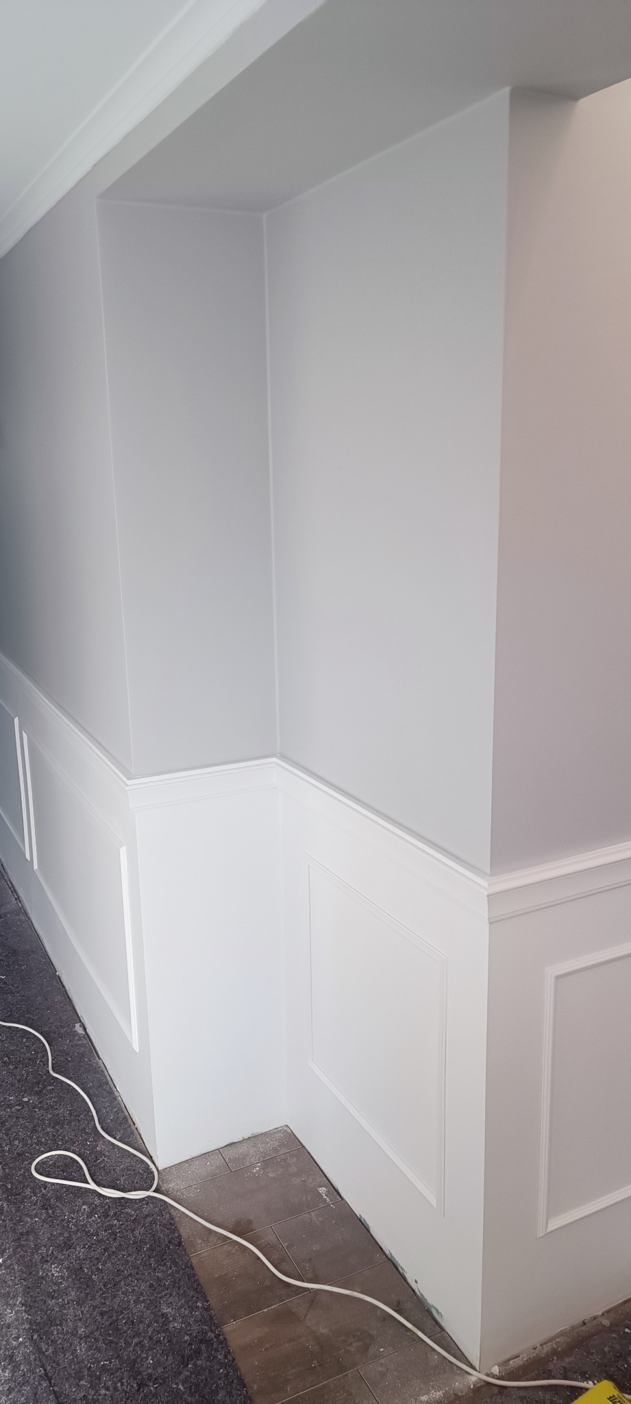 Świeżo pomalowany narożnik pokoju z dekoracyjną białą boazerią angielską i listwami przypodłogowymi, zabezpieczona podłoga i kabel zasilający na posadzce.