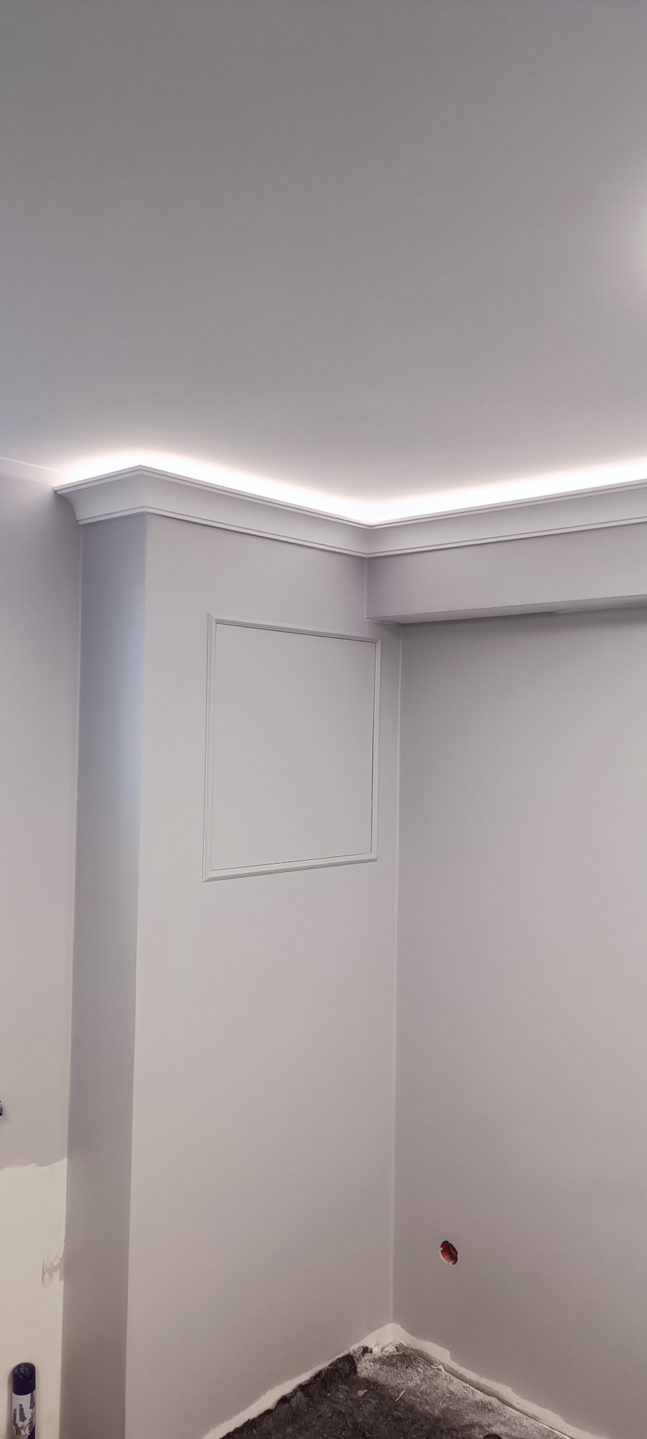 Szare wnętrze po szpachlowaniu, widoczne listwy przysufitowe z oświetleniem LED, otwór rewizyjny w ścianie, ślady po pracach na podłodze i niedomalowany fragment narożnika.