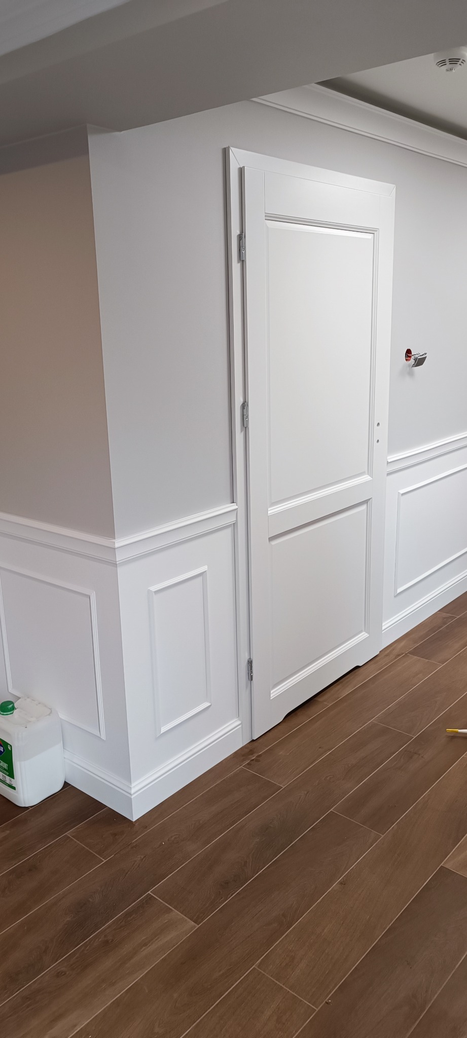 Białe, dwuskrzydłowe drzwi wewnętrzne zamontowane w ścianie z dekoracyjną sztukaterią, widoczne brązowe panele podłogowe i pojemnik z płynem do czyszczenia w rogu.