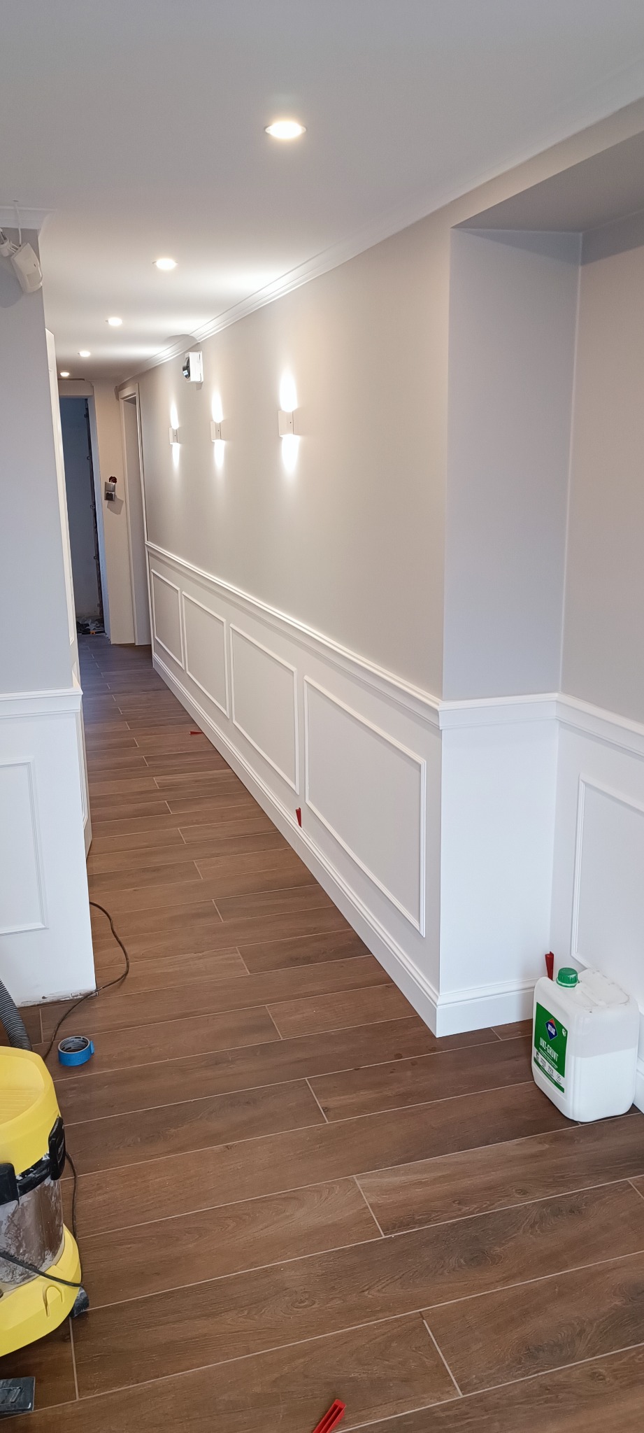 Korytarz po remoncie z drewnopodobną podłogą, panelami ściennymi i nowoczesnym oświetleniem, widoczny odkurzacz i pojemnik z gruntem.