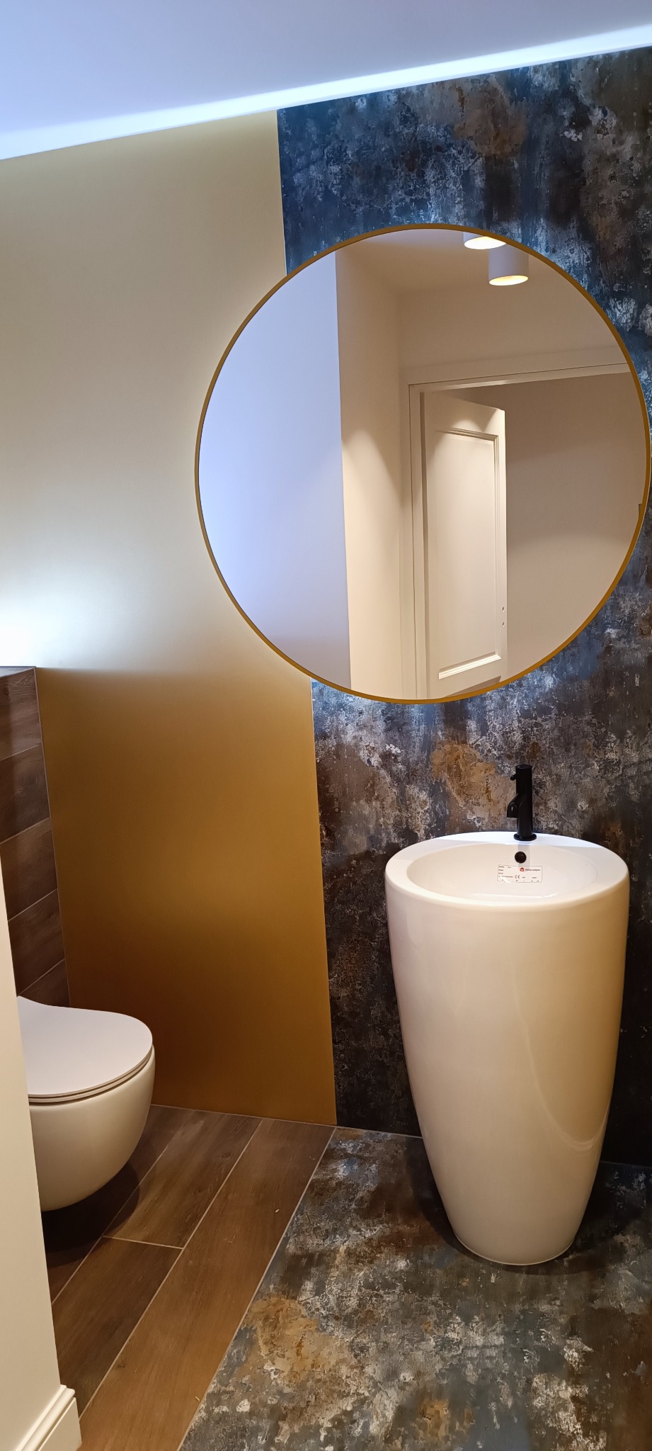 Nowoczesna łazienka z umywalką na postumencie, okrągłym lustrem z podświetleniem i płytkami imitującymi rdzę, widok na otwarte drzwi.