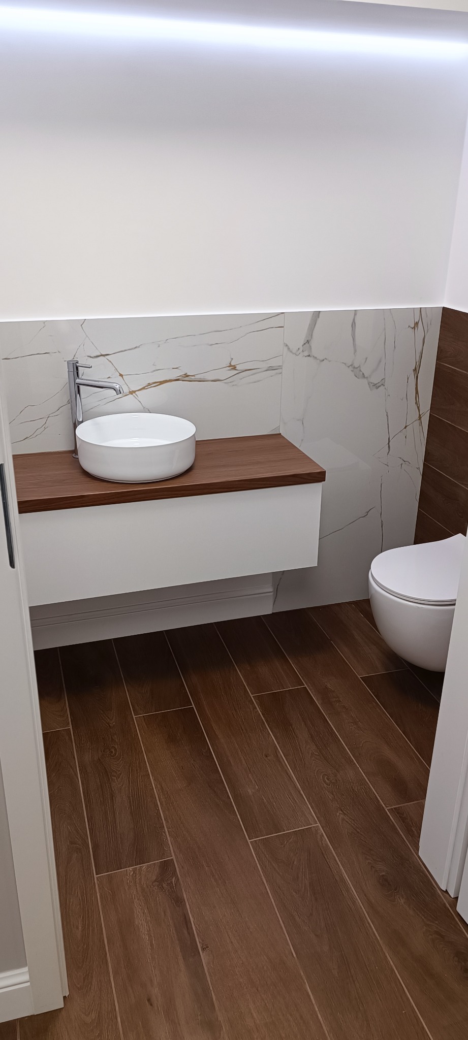 Nowoczesna łazienka z okrągłą umywalką nablatową, blatem z ciemnego drewna, białymi płytkami imitującymi marmur z złotymi żyłami oraz brązowymi płytkami podłogowymi imitującymi drewno, oświetleniem...