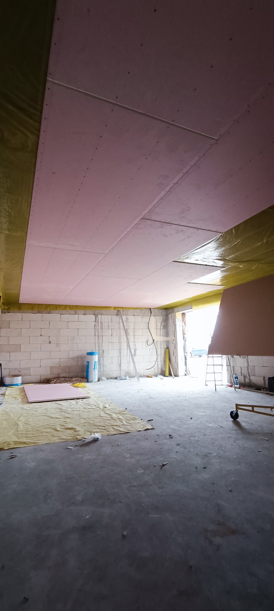 Montaż płyt kartonowo-gipsowych na suficie w surowym pomieszczeniu, widoczne profile, izolacja i przygotowane płyty na podłodze.