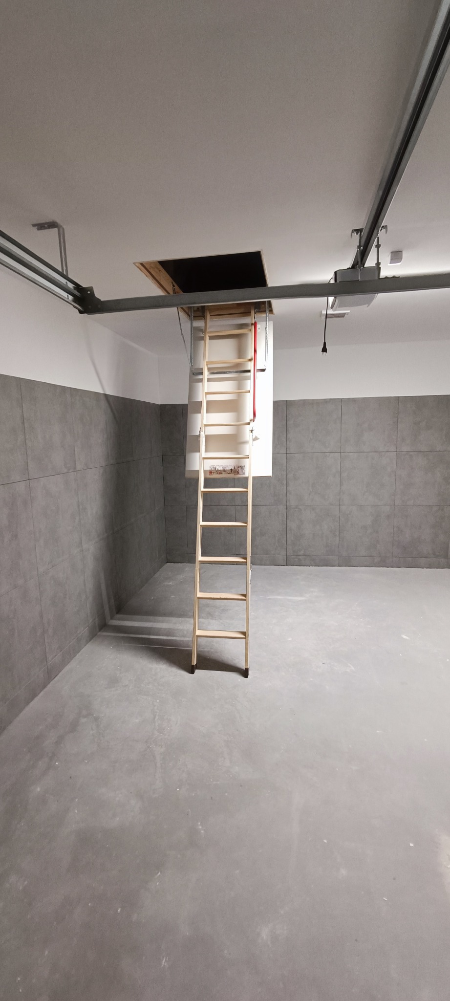 Zamontowane drewniane schody strychowe prowadzące do otworu w suficie w pomieszczeniu z szarymi płytkami na ścianie i betonową podłogą.