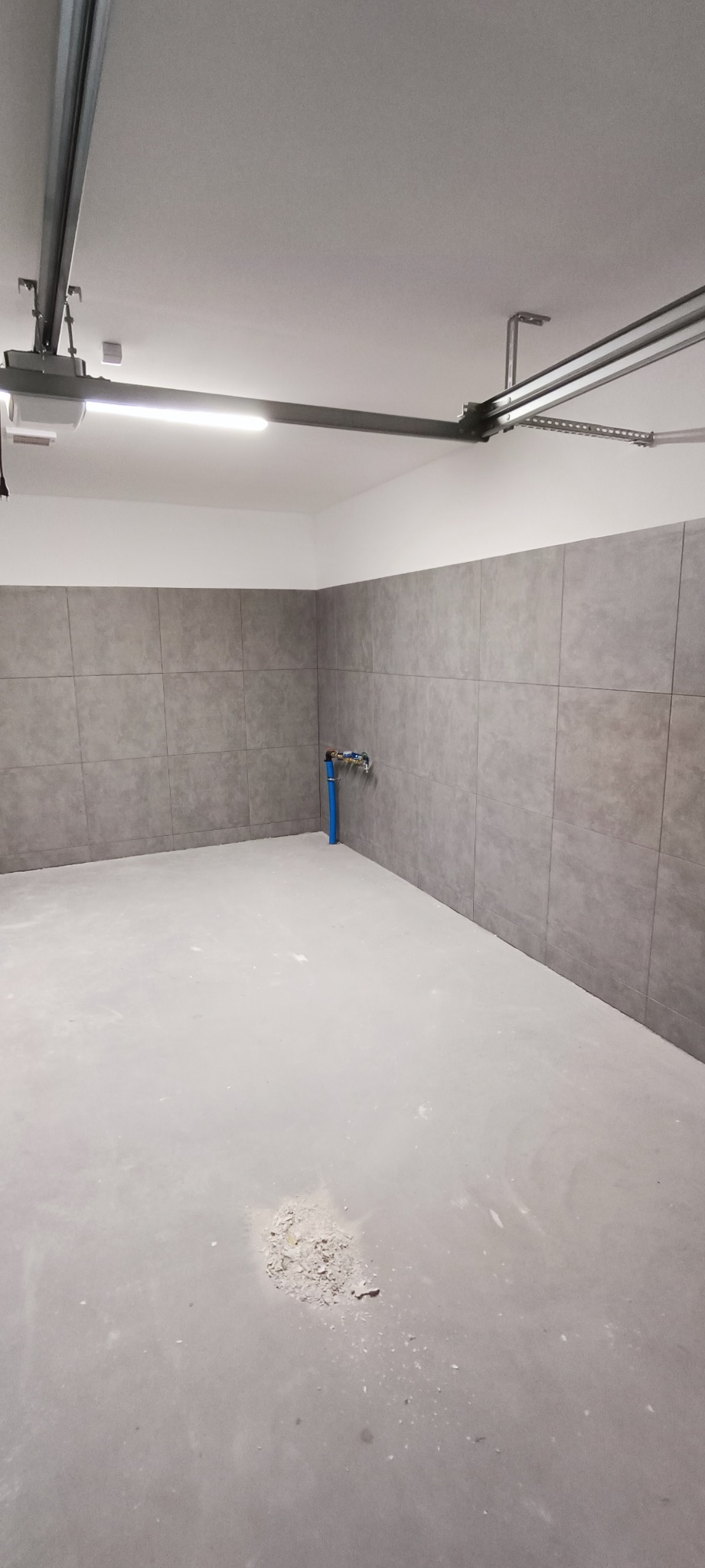 Wnętrze łazienki w Szczecinie po położeniu szarych płytek na ścianach, widoczna niebieska rura z zaworem i pozostałości po pracach na szarej podłodze.