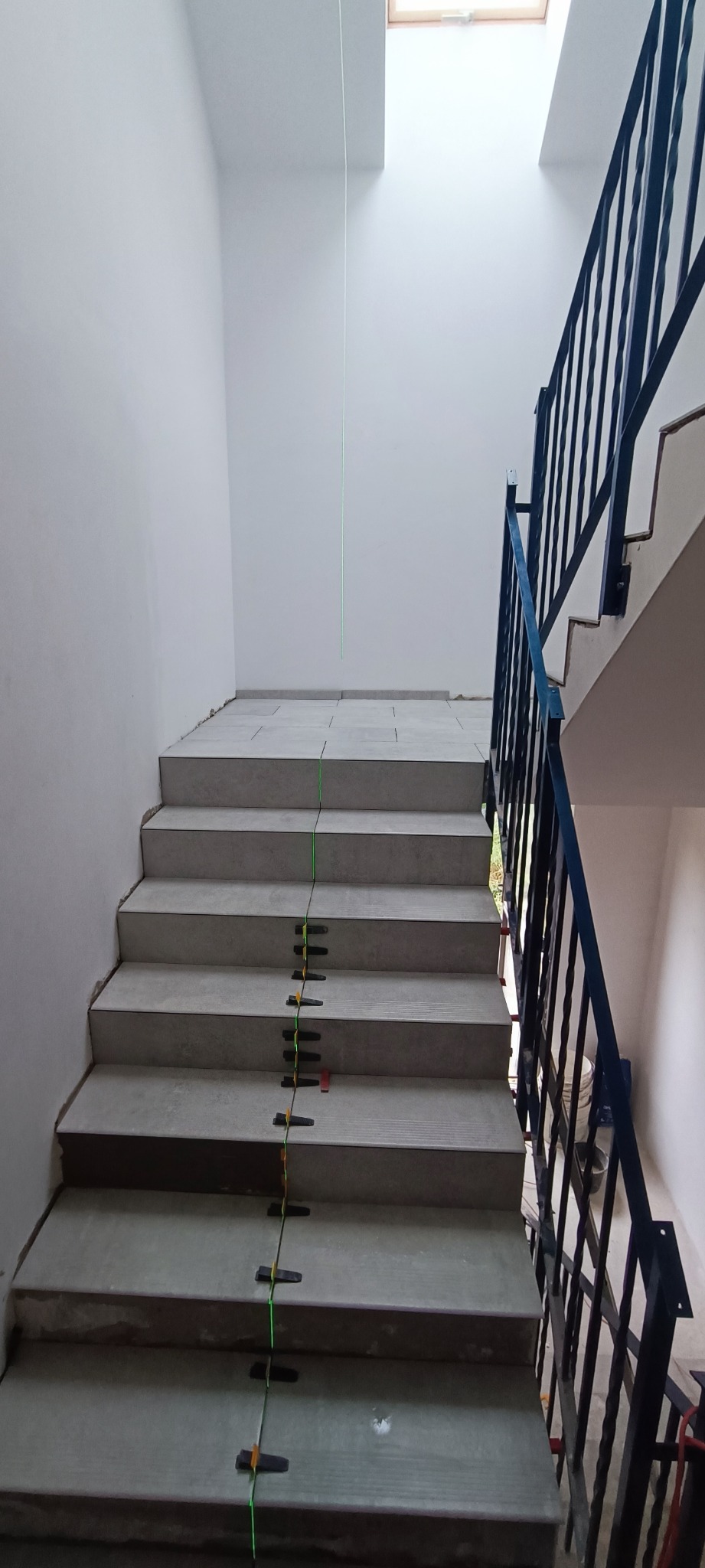 Schody wyłożone szarymi płytkami, poziomowane laserowo z użyciem czarnych klipsów dystansowych i czerwonego krzyżyka, widoczna czarna balustrada i okno dachowe.
