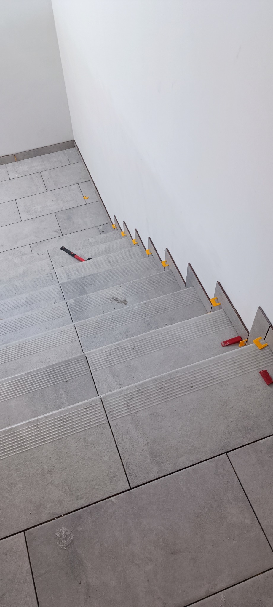 Schody wyłożone szarymi płytkami w trakcie prac wykończeniowych, z użyciem klipsów poziomujących i młotka gumowego.