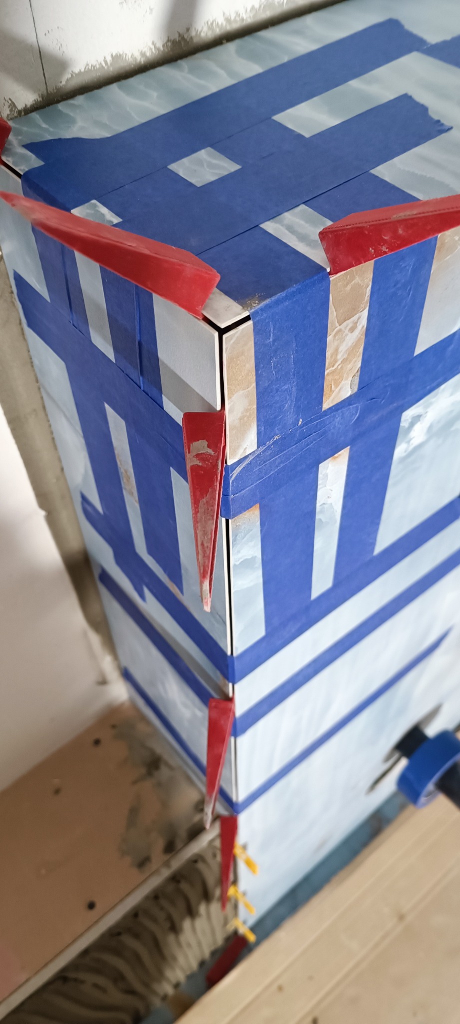 Kąt narożny obłożony błyszczącymi, wzorzystymi płytkami, zabezpieczony niebieską taśmą malarską i czerwonymi klinami dystansowymi, widoczne żółte krzyżyki dystansowe i fragment drewnianej konstrukcji.