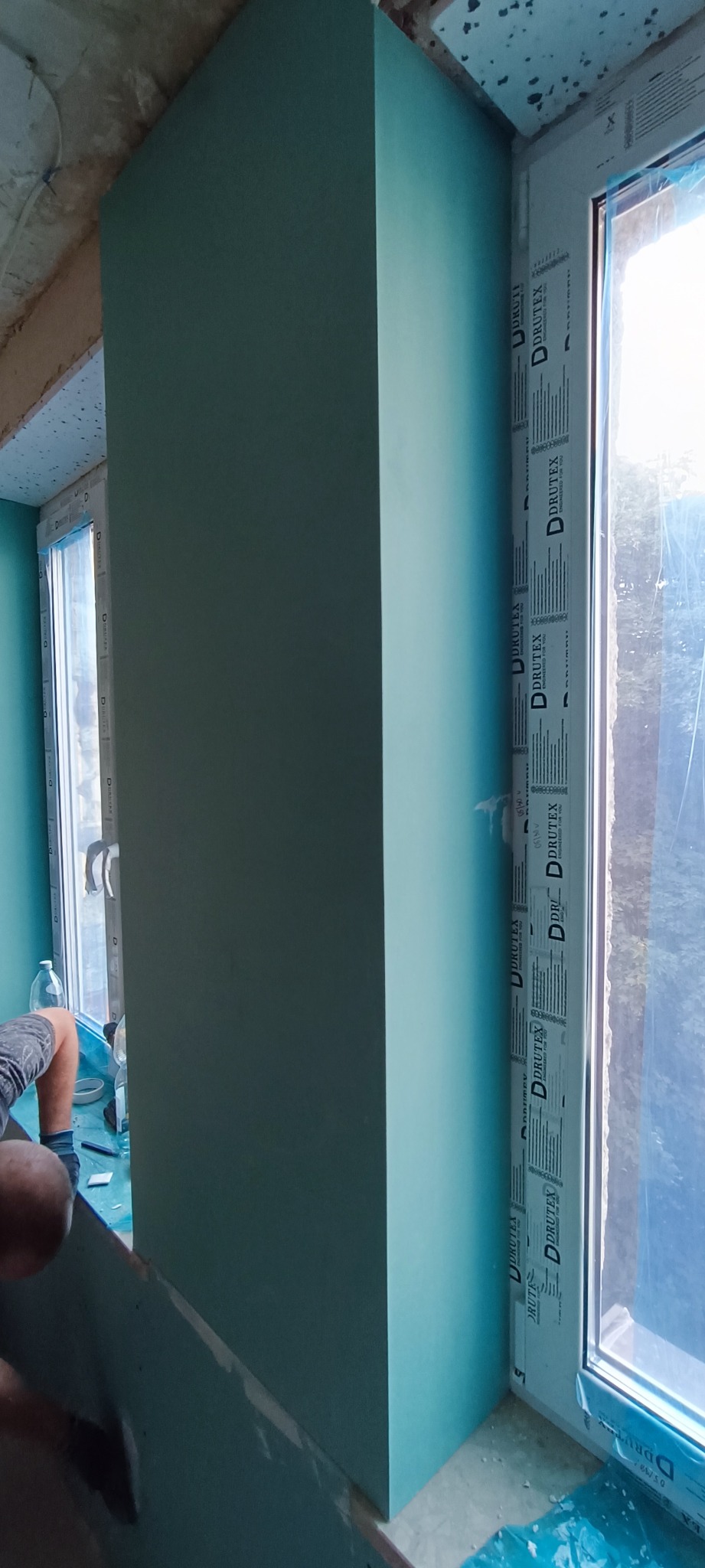 Montaż pionowej konstrukcji z płyty gipsowo-kartonowej przy oknie z widocznym logo producenta, fragment pracownika w tle, zabezpieczona folią podłoga.