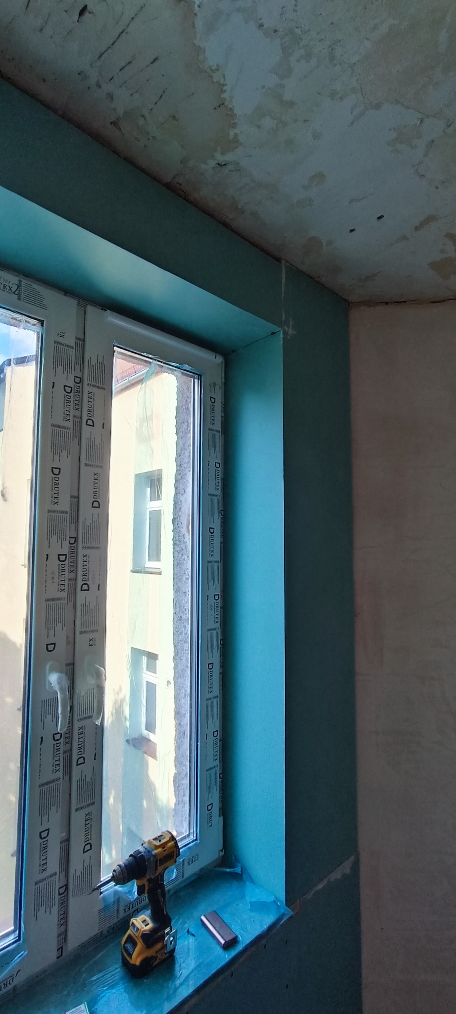 Montaż zielonej płyty kartonowo-gipsowej wokół okna, widoczna wiertarka akumulatorowa na parapecie, surowy sufit w tle.