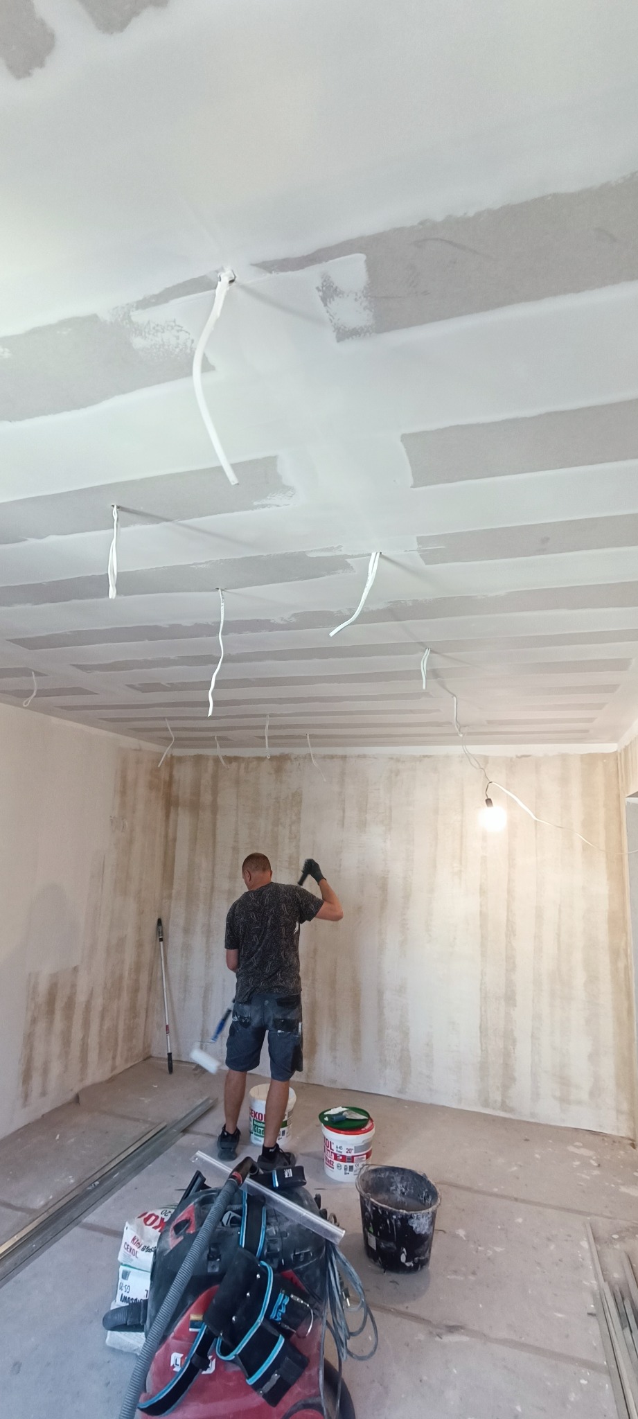Mężczyzna w krótkich spodenkach i czarnej koszulce maluje ścianę wałkiem w pomieszczeniu z płytami kartonowo-gipsowymi na suficie, widoczne przewody elektryczne, wiaderka z farbą i odkurzacz...
