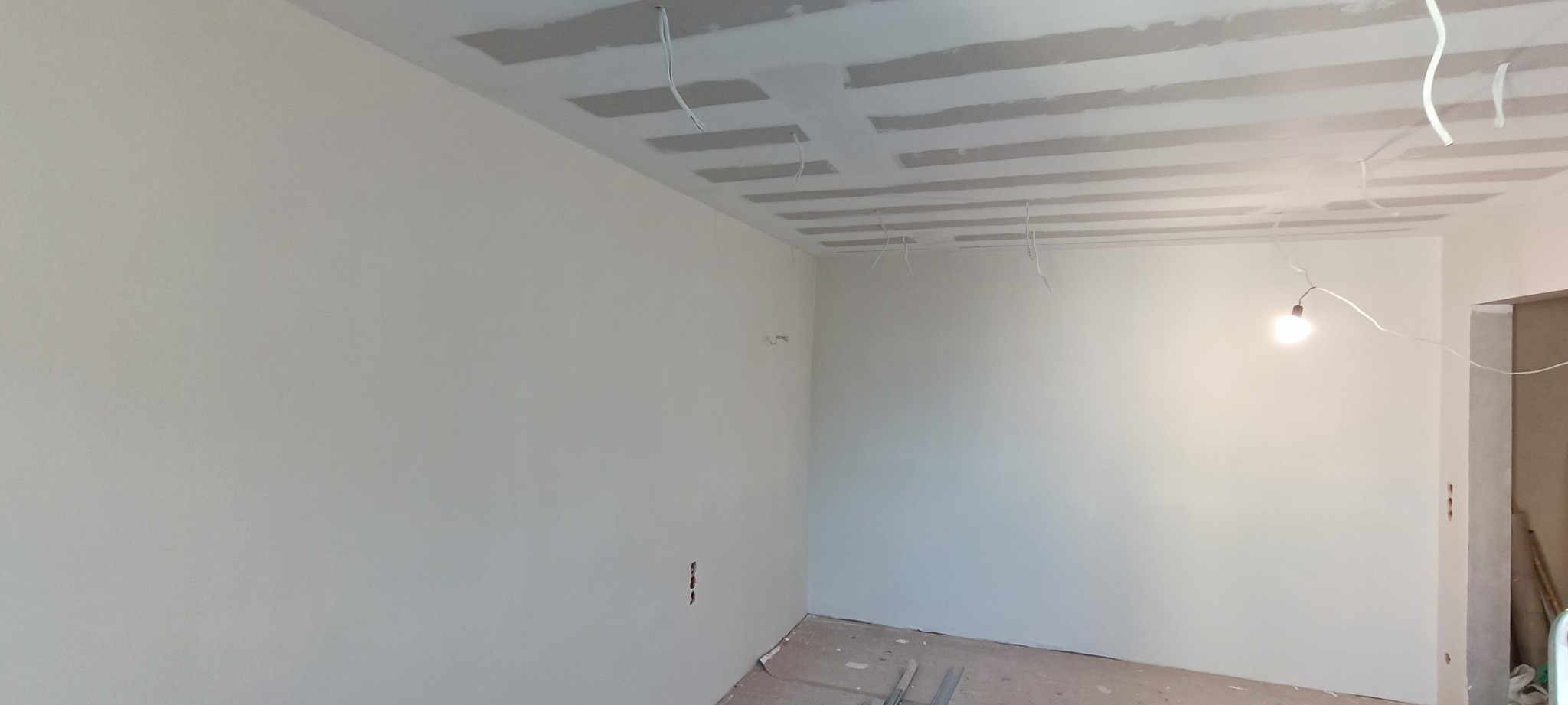 Pokój w trakcie prac wykończeniowych: widoczne szpachlowane ściany, sufit z konstrukcją pod płyty gipsowe, wystające przewody elektryczne i prowizoryczne oświetlenie.