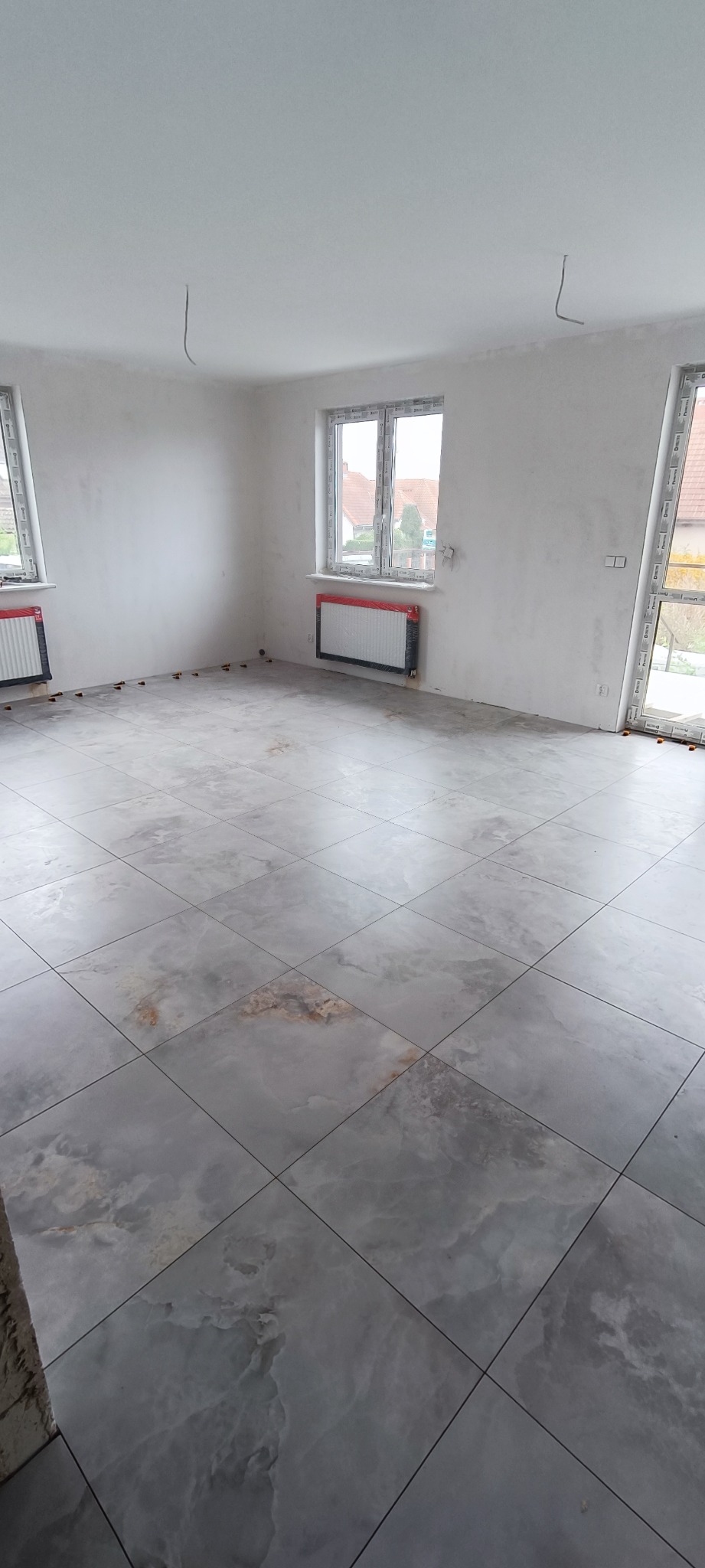 Salon z nowo położoną, szarą, marmurkową podłogą z płytek, zabezpieczone grzejniki folią, widoczne odstające kable ze ściany.