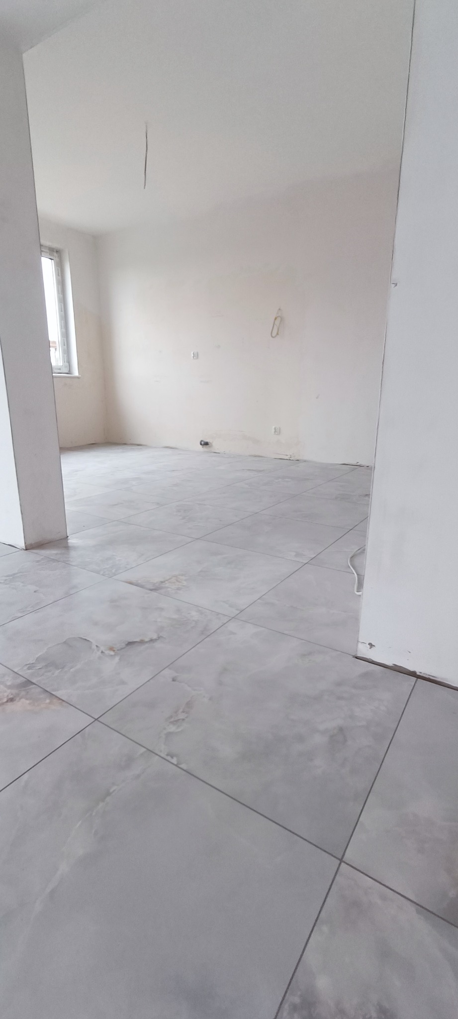 Wyłożona szarymi, marmurkowymi płytkami podłoga w surowym wnętrzu z białymi ścianami i widoczną instalacją elektryczną.