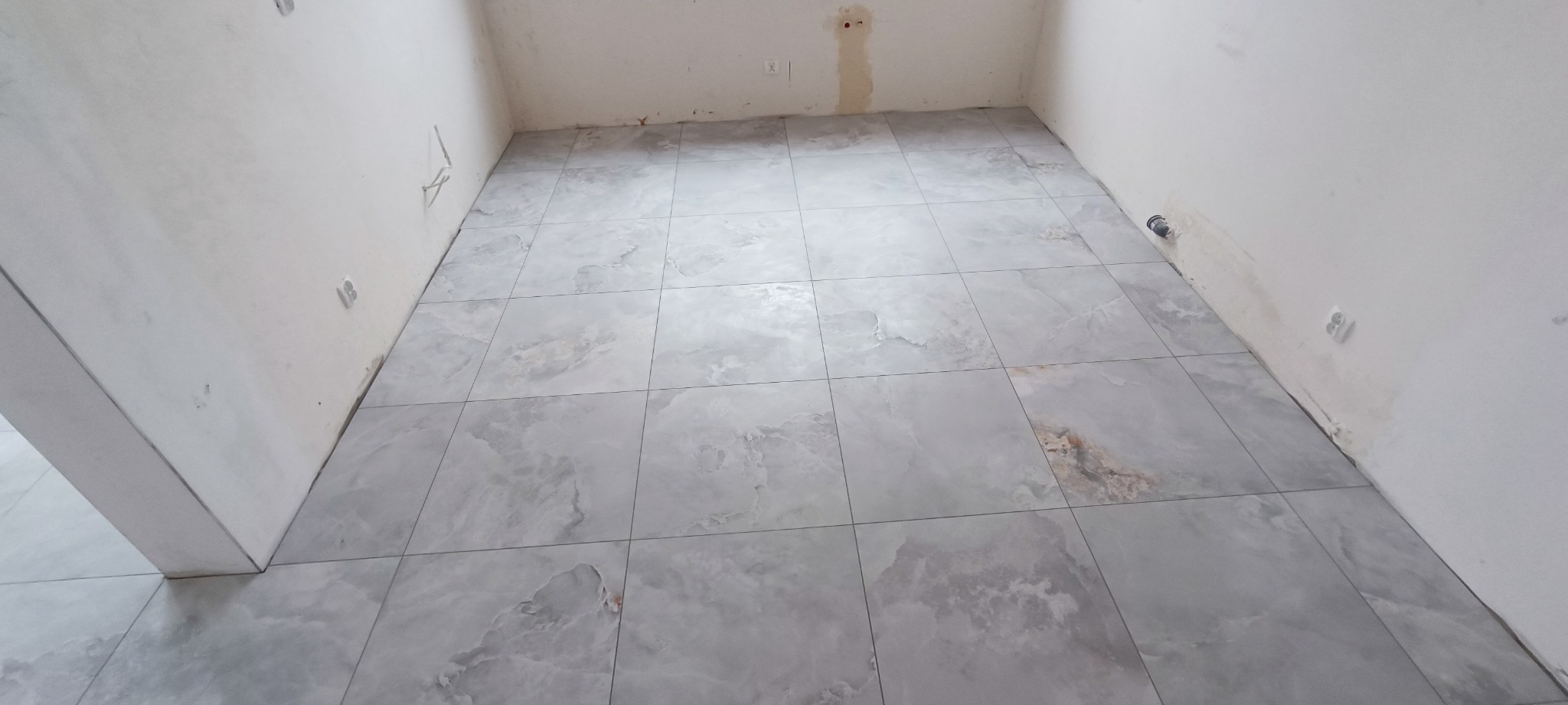 Ułożona szara, marmurkowana podłoga z płytek w surowym pomieszczeniu z widocznymi śladami po instalacjach elektrycznych na ścianach.
