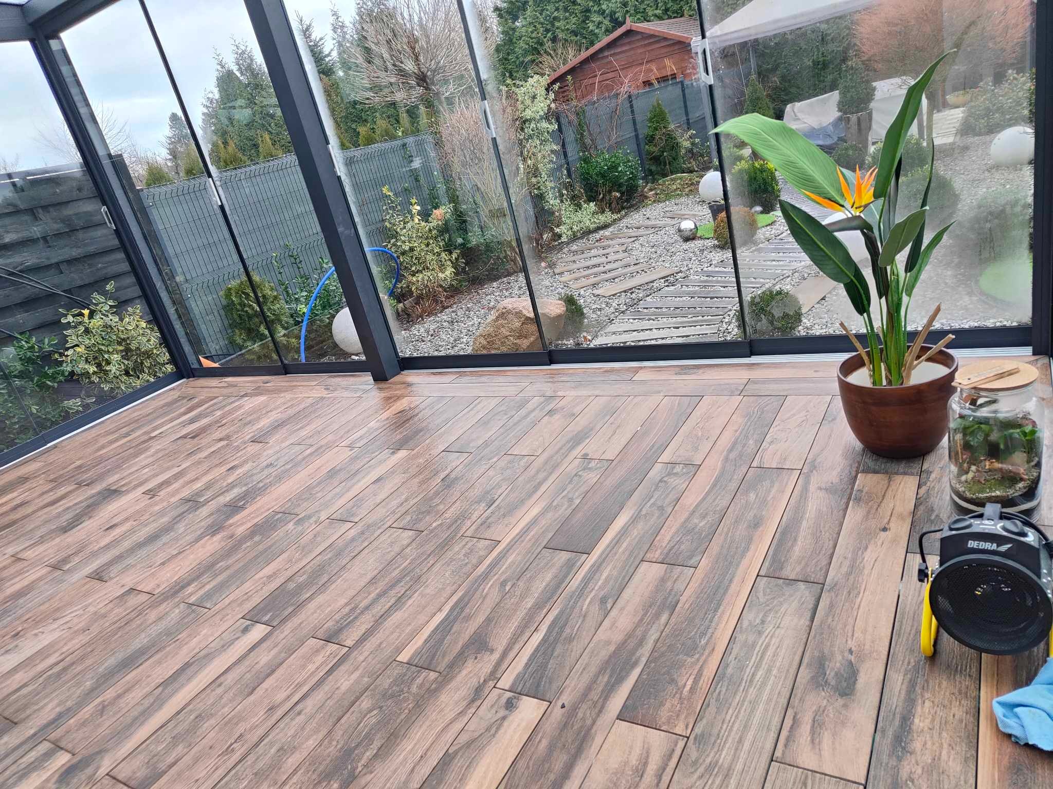 Drewnopodobna podłoga w oranżerii z widokiem na ogród, w rogu stoi egzotyczny kwiat w doniczce oraz nagrzewnica Dedra.