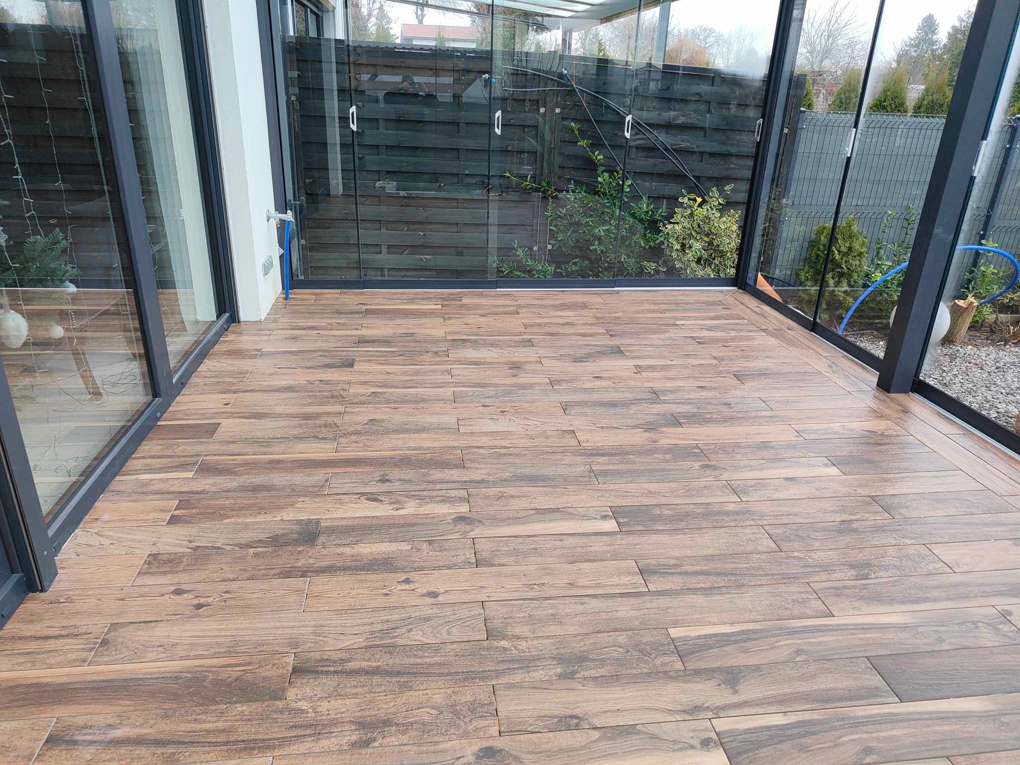 Drewnopodobne płytki podłogowe ułożone w oranżerii z przeszklonymi ścianami i widokiem na ogród.