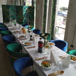 Uroczysta aranżacja stołu z białym obrusem, złotymi sztućcami, daniami na talerzach i napojami, otoczona zielono-niebieskimi krzesłami, z dekoracyjnym parawanem w tle i roślinami pnącymi się po...