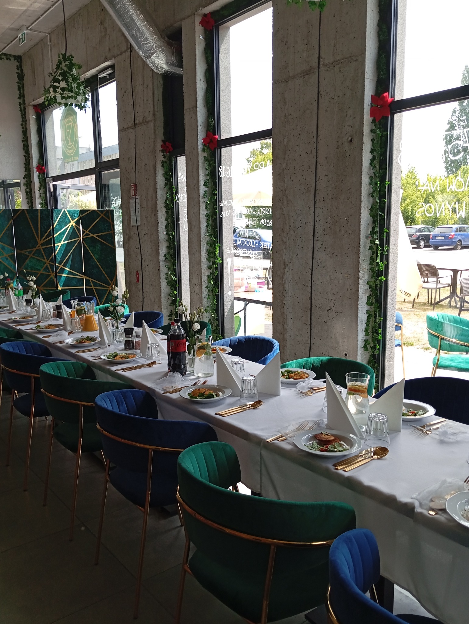 Uroczysty stół z białym obrusem, zastawą stołową w kolorze złota, szklankami, napojami i daniami, otoczony krzesłami w odcieniach zieleni i granatu, w przestronnym wnętrzu z betonowymi kolumnami...