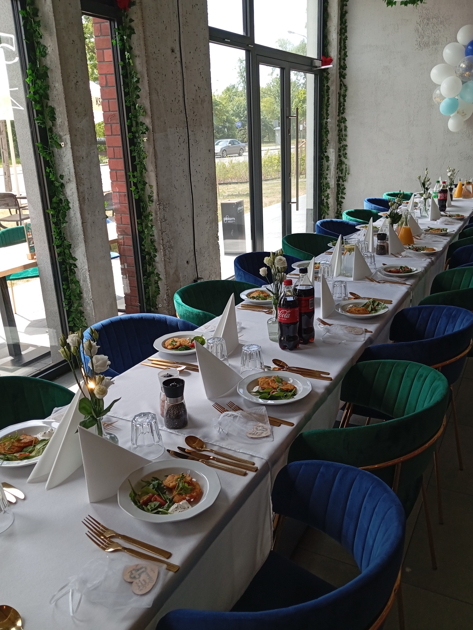 Uroczysty stół z białym obrusem, nakryty złotymi sztućcami i talerzami z sałatkami, otoczony welurowymi krzesłami w odcieniach zieleni i granatu, dekorowany kompozycjami kwiatowymi i girlandami...