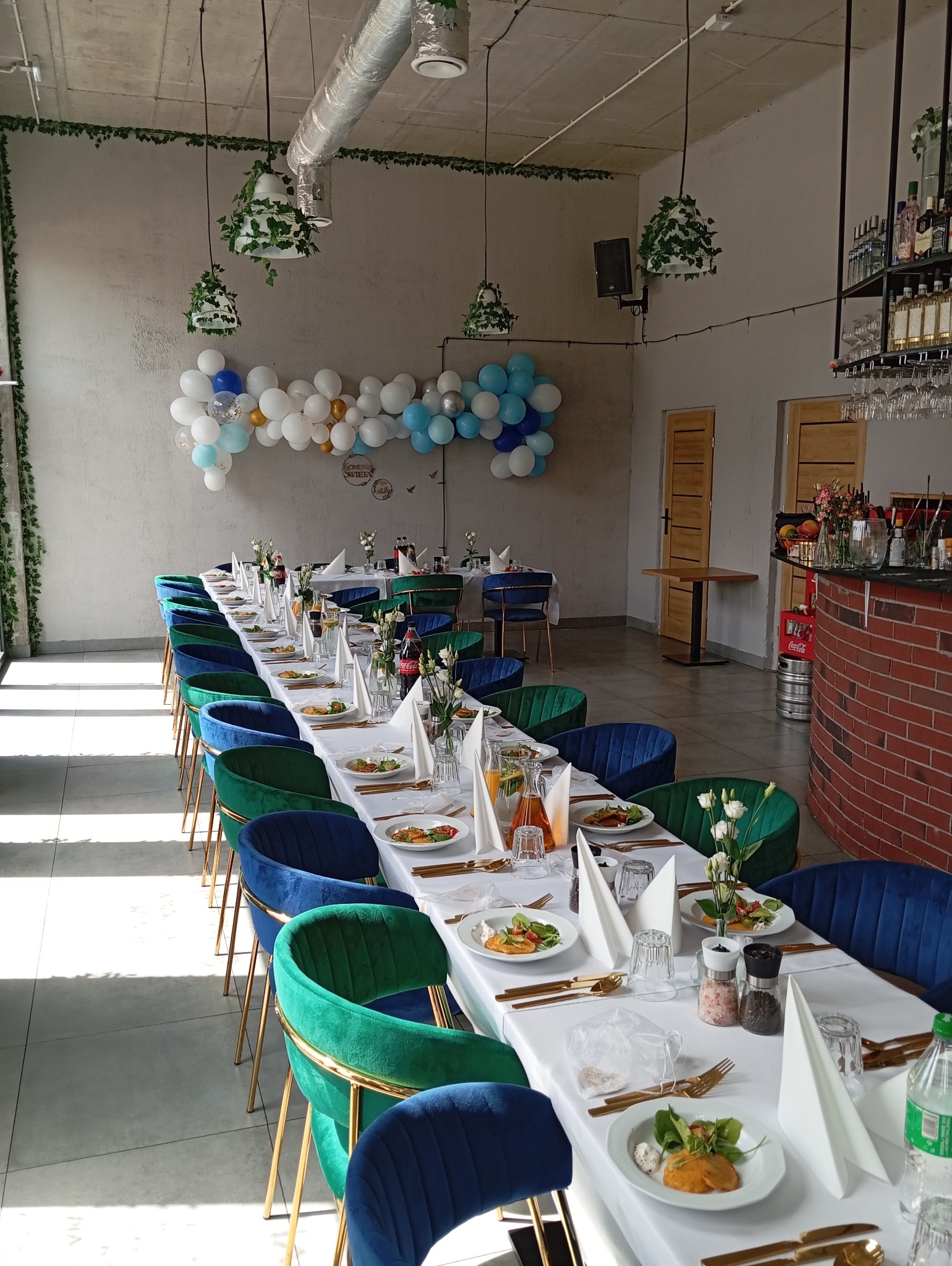 Uroczysta sala z długim stołem nakrytym białym obrusem, złotymi sztućcami, talerzami z przystawkami i dekoracjami kwiatowymi, krzesła obite aksamitem w kolorach zieleni i granatu, ściana ozdobiona...