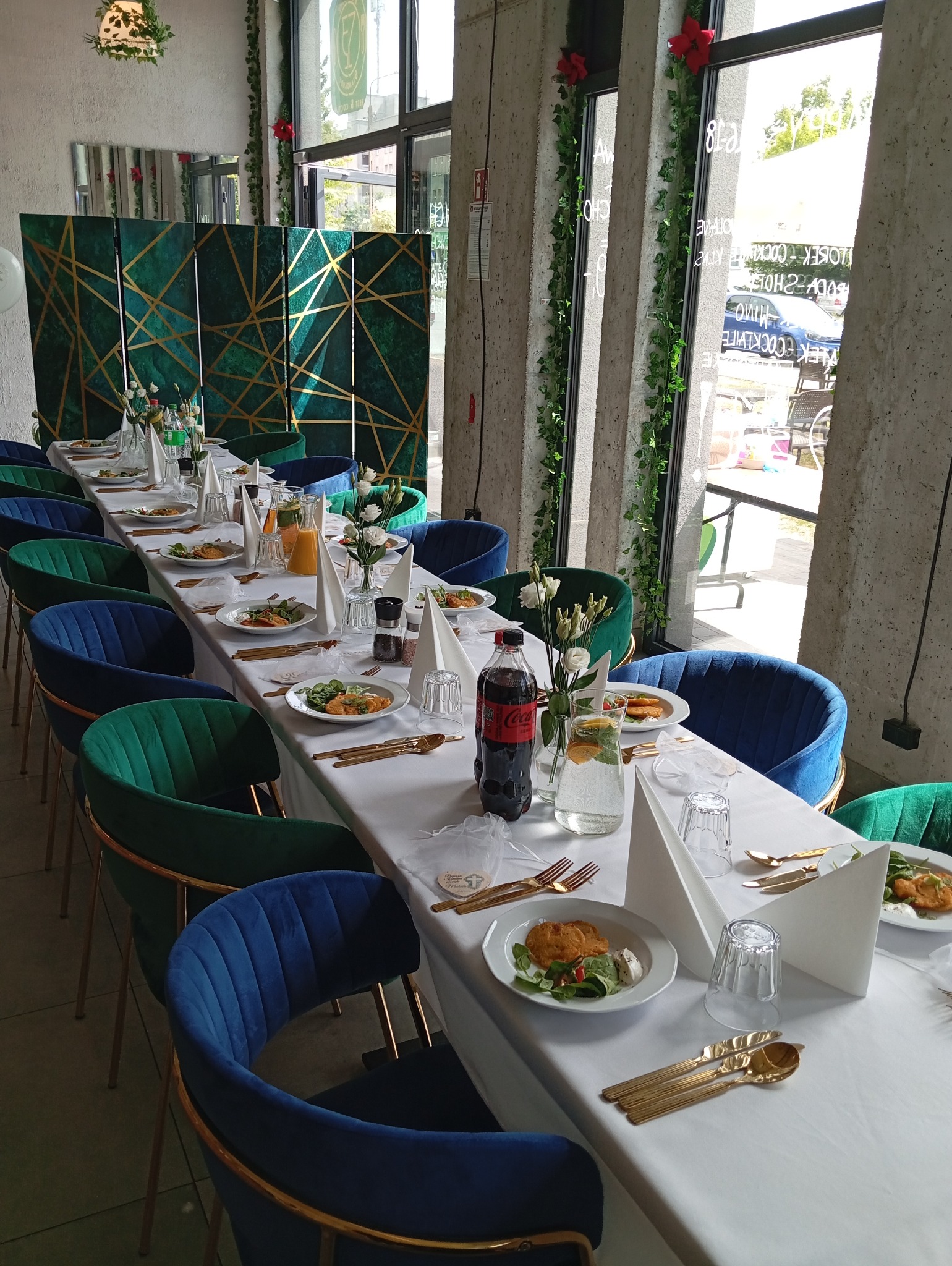 Uroczysta aranżacja stołu z białym obrusem, złotymi sztućcami, daniami na talerzach i napojami, otoczona zielono-niebieskimi krzesłami, z dekoracyjnym parawanem w tle i roślinami pnącymi się po...