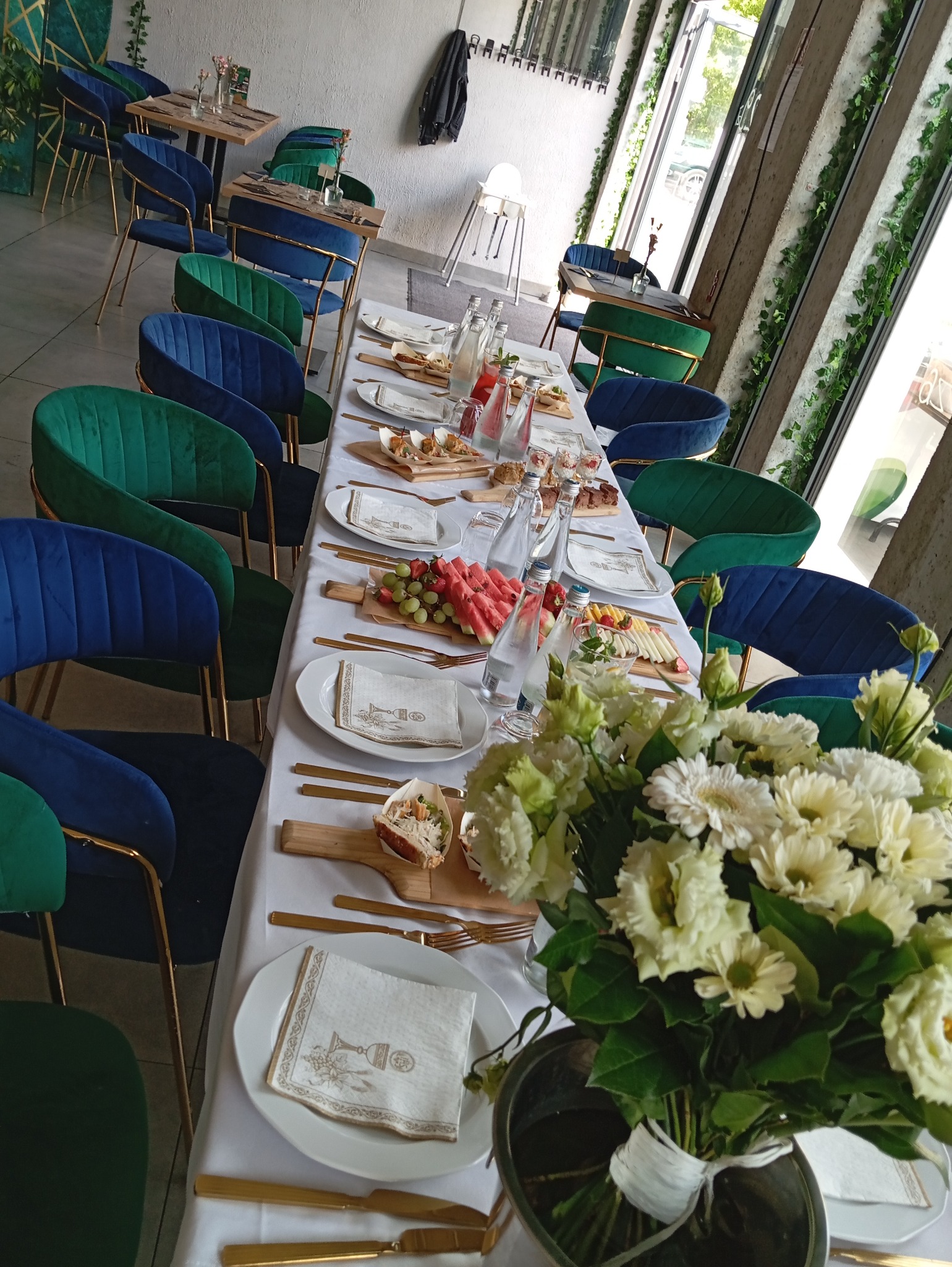 Długi stół z białym obrusem, zastawiony złotymi sztućcami, talerzami z serwetkami z motywem kielicha, przekąskami, owocami (arbuz, truskawki, winogrona) i wodą mineralną; w tle zielono-niebieskie...