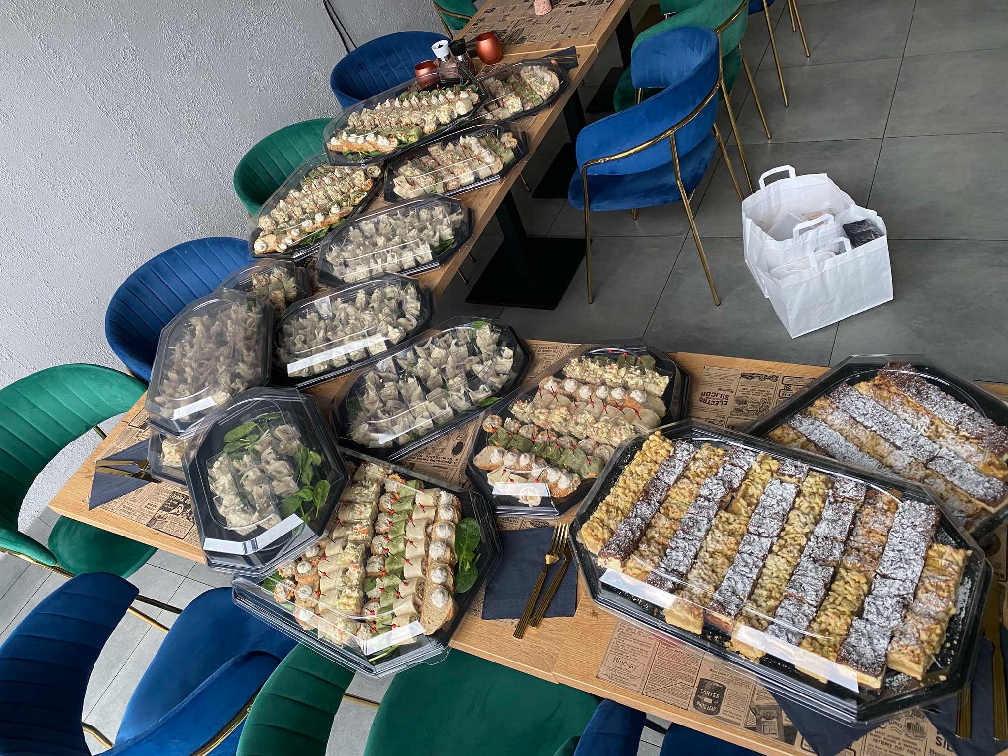 Stół z drewnianym blatem pokryty gazetami, na którym ustawiono plastikowe pojemniki z różnorodnymi przekąskami, w tle krzesła w kolorach niebieskim i zielonym.