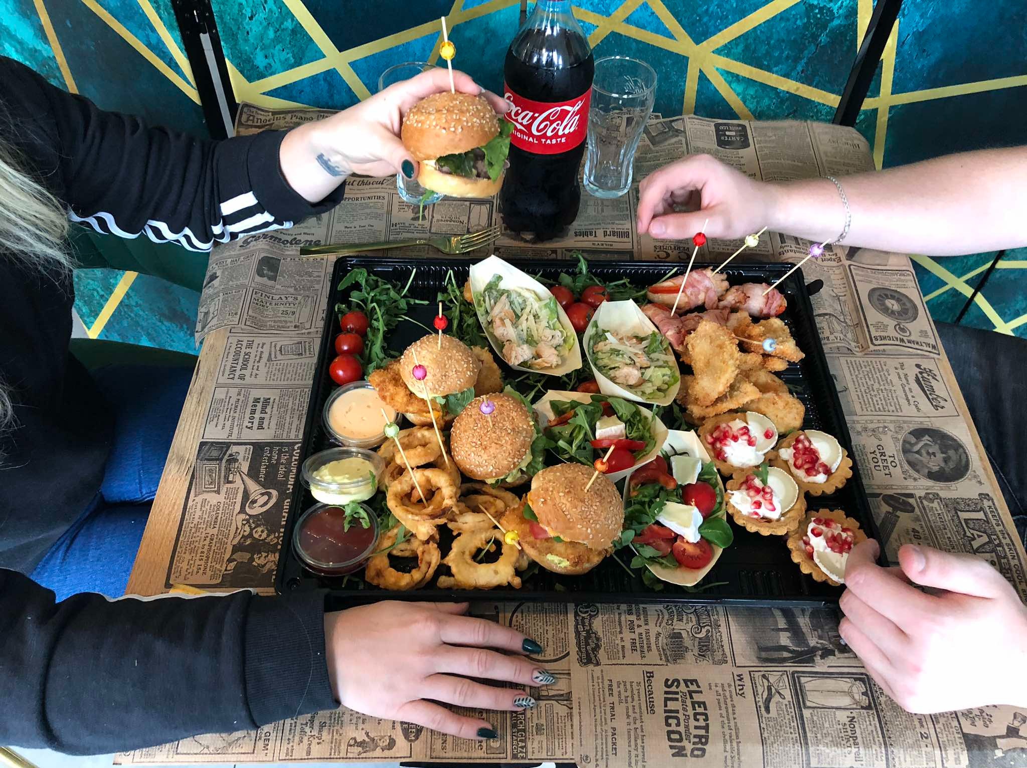 Czarny cateringowy półmisek z mini burgerami, tartaletkami z serem i granatem, krążkami cebulowymi, sałatkami w łódeczkach i przekąskami owiniętymi boczkiem, trzymany przez dwie osoby na stole...
