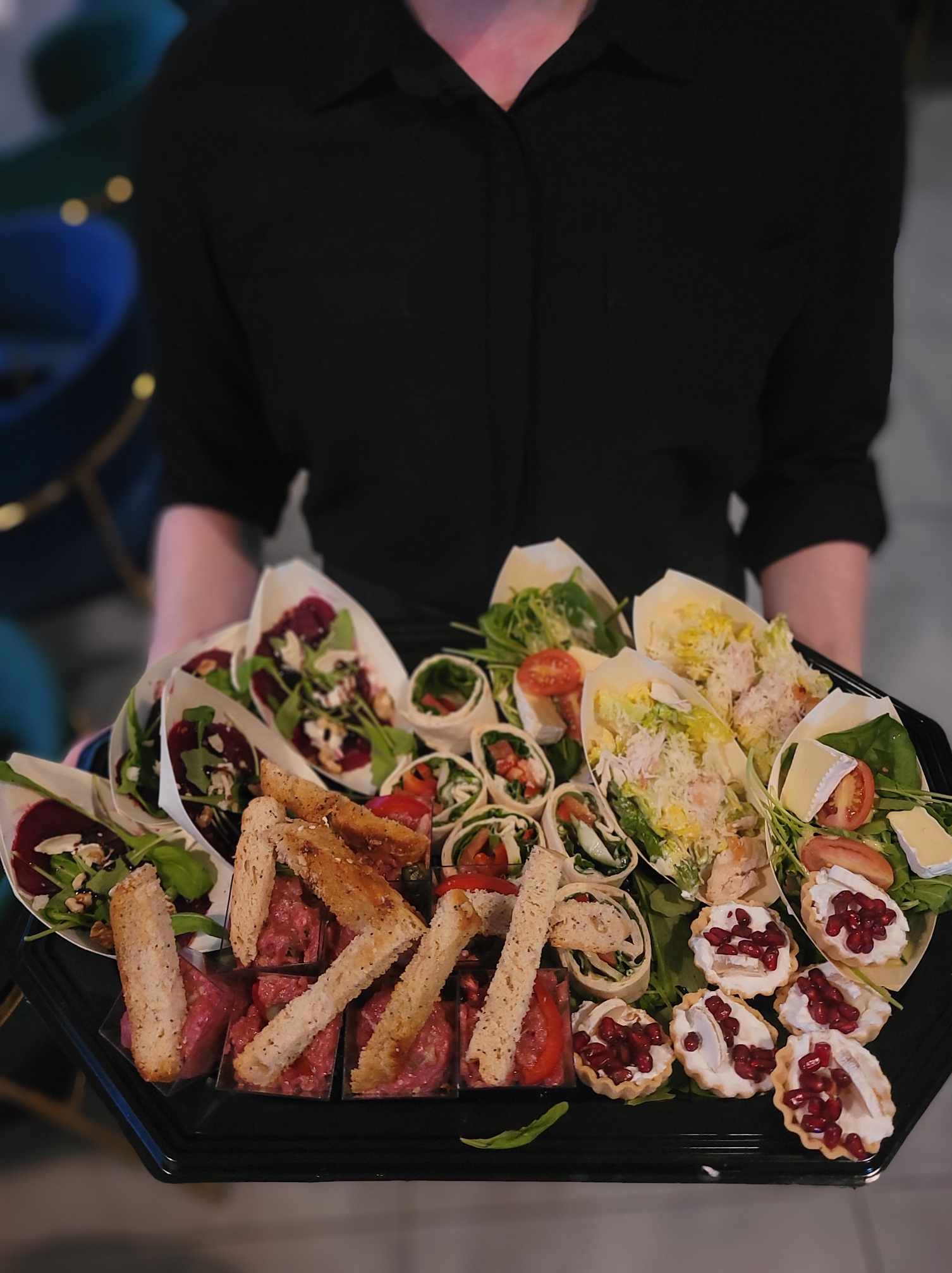 Osoba w czarnej koszuli trzyma czarną tacę z różnorodnymi przekąskami cateringowymi: tartaletki z granatem, sałatki w papierowych łódeczkach, roladki z warzywami i mięsem, tatar wołowy z grzankami.