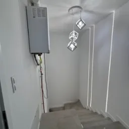 Klatka schodowa z szarymi płytkami, oświetlona nowoczesną lampą wiszącą z geometrycznymi kloszami i pionowymi pasami światła w ścianie, z widocznym piecem gazowym na ścianie.